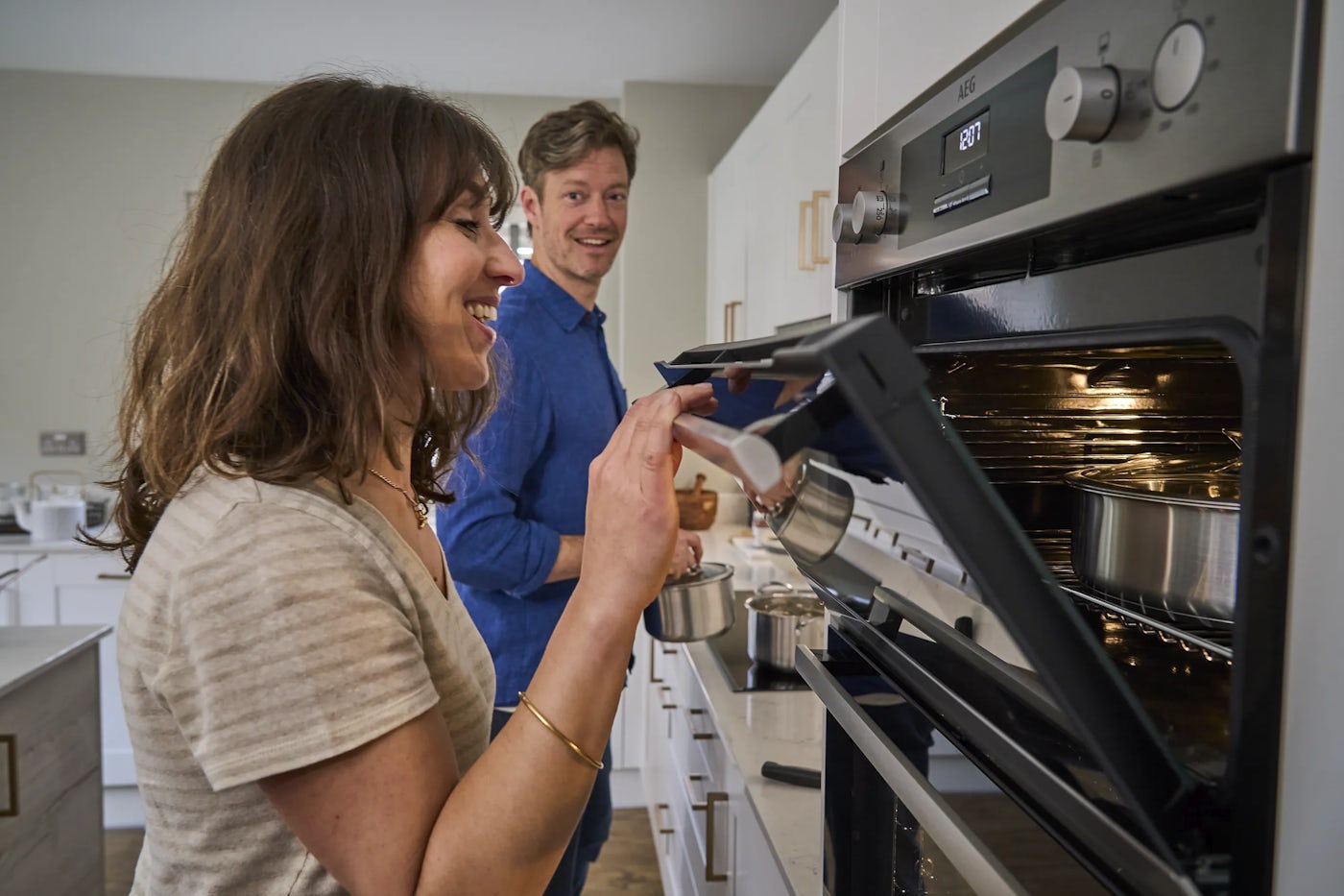 Two people in a modern kitchen. A woman is opening an oven door, revealing a round baking pan inside, while the man stands behind holding a pot. White cabinets and stainless steel appliances frame the scene.