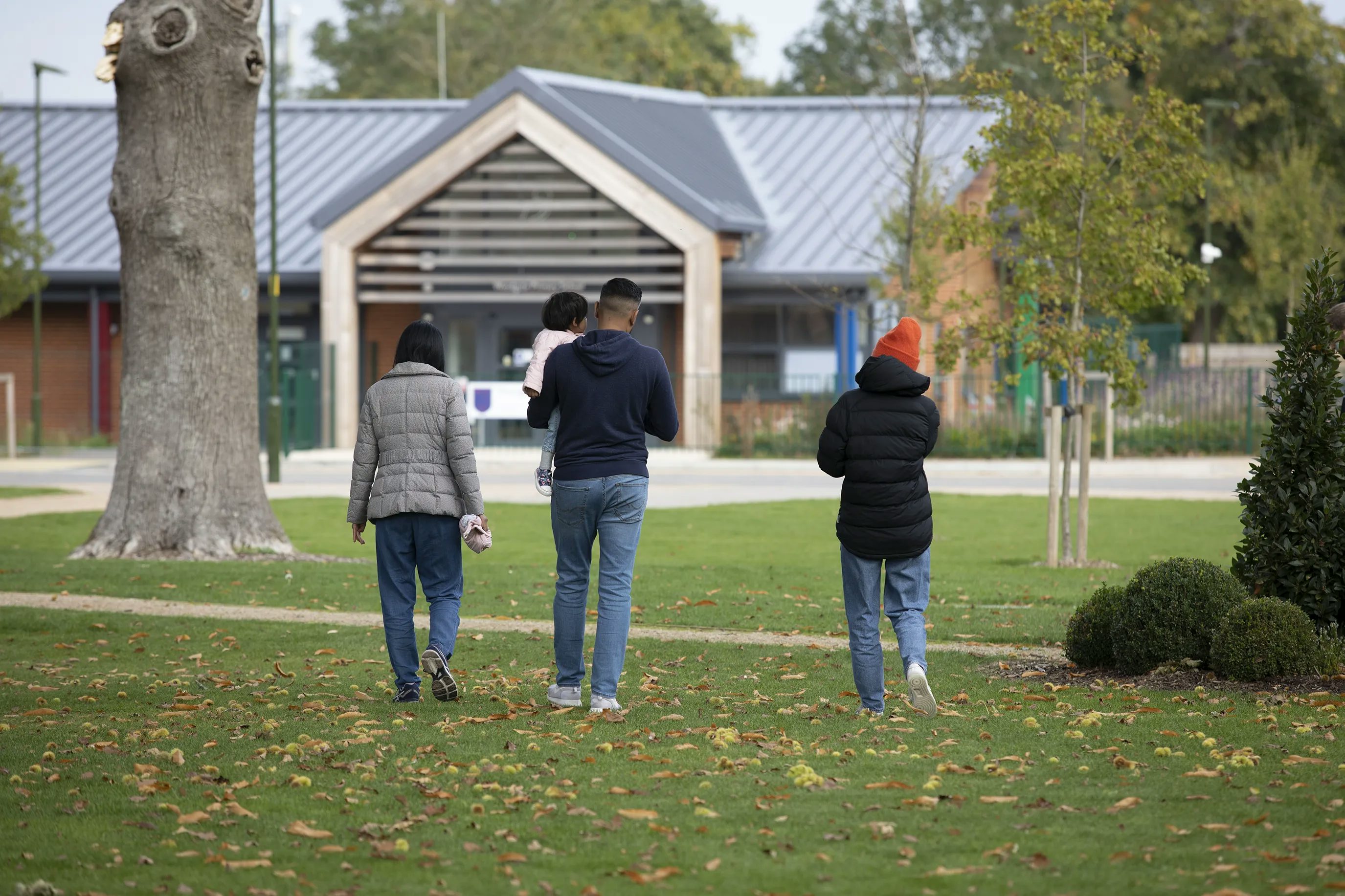 Three adults walk through a park with green grass, fallen leaves, trees, and bushes. One adult carries a child as they stroll together. A modern building with a triangular roof and large windows stands in the background. The scene captures a casual, everyday moment outdoors.