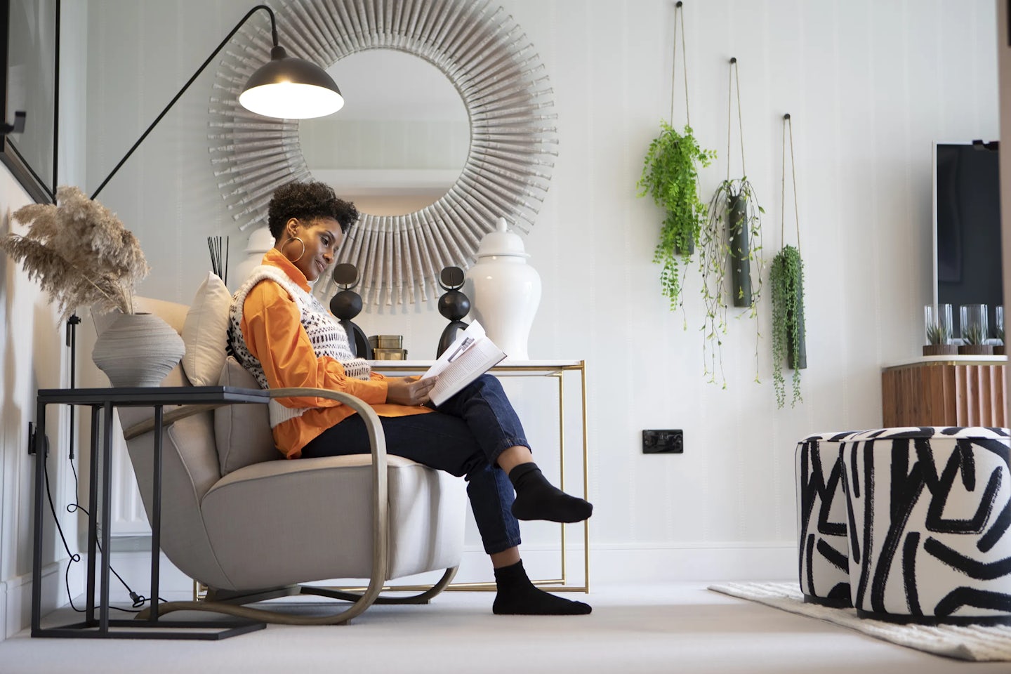 A woman sits in a modern living room, reading a book or magazine in a light-colored armchair with a metal frame. The room has contemporary decor, including a large circular mirror with a textured frame, hanging plants, and a geometric-patterned ottoman. A wooden cabinet with a television sits against the wall, with a floor lamp nearby. A side table holds decorative items, and a vase with pampas grass adds a natural touch.