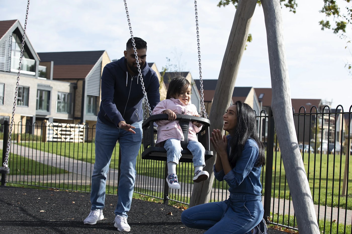 A person pushes a child on a swing at a playground, while another person kneels nearby, possibly interacting with the child. The playground is in a residential area, with modern houses visible in the background. The scene captures a family moment of outdoor activity.