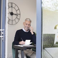 A man sits at a desk in a home office or study, viewed through a glass door. He holds a phone to his ear and writes on a notepad. Behind him, a large wall clock with Roman numerals is visible. The desk has a lamp, a container with pencils, and scattered papers.