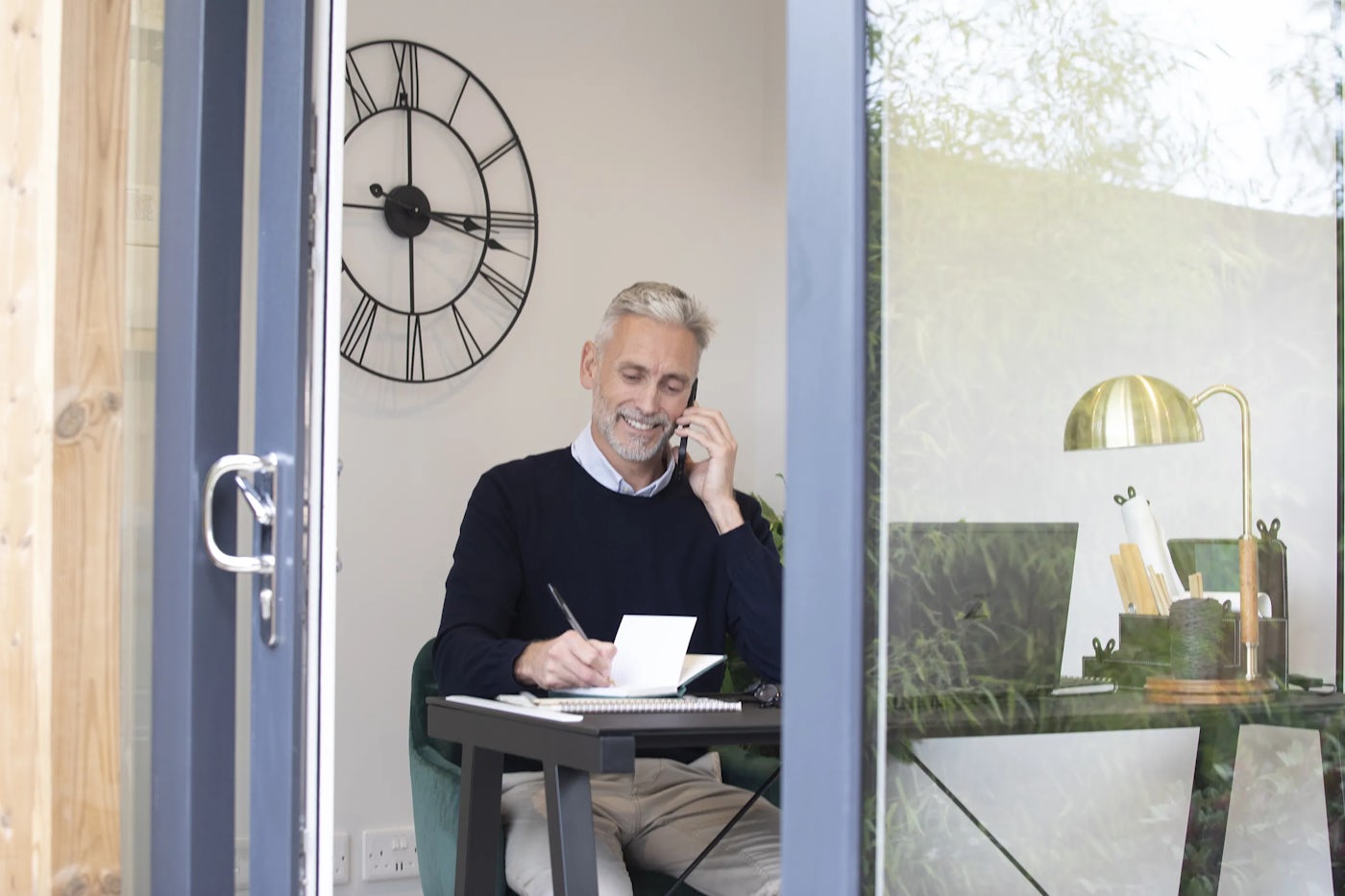 A man sits at a desk in a home office or study, viewed through a glass door. He holds a phone to his ear and writes on a notepad. Behind him, a large wall clock with Roman numerals is visible. The desk has a lamp, a container with pencils, and scattered papers.