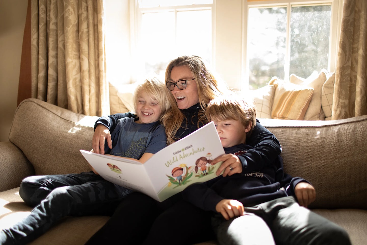 The image shows a woman sitting on a couch with two young children. She is reading a book titled Eddie and Ellies Wild Adventure to them. The room is well-lit, with large windows and curtains in the background. The book cover features illustrations of children and animals, suggesting it is a children's story.