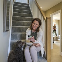 A woman sits on a staircase, holding a smartphone while a dog sits beside her, looking up. The staircase has grey carpet and wooden railings. In the background, a man stands in a room, holding a mug, with a hallway leading to a white door and a small table with a plant.