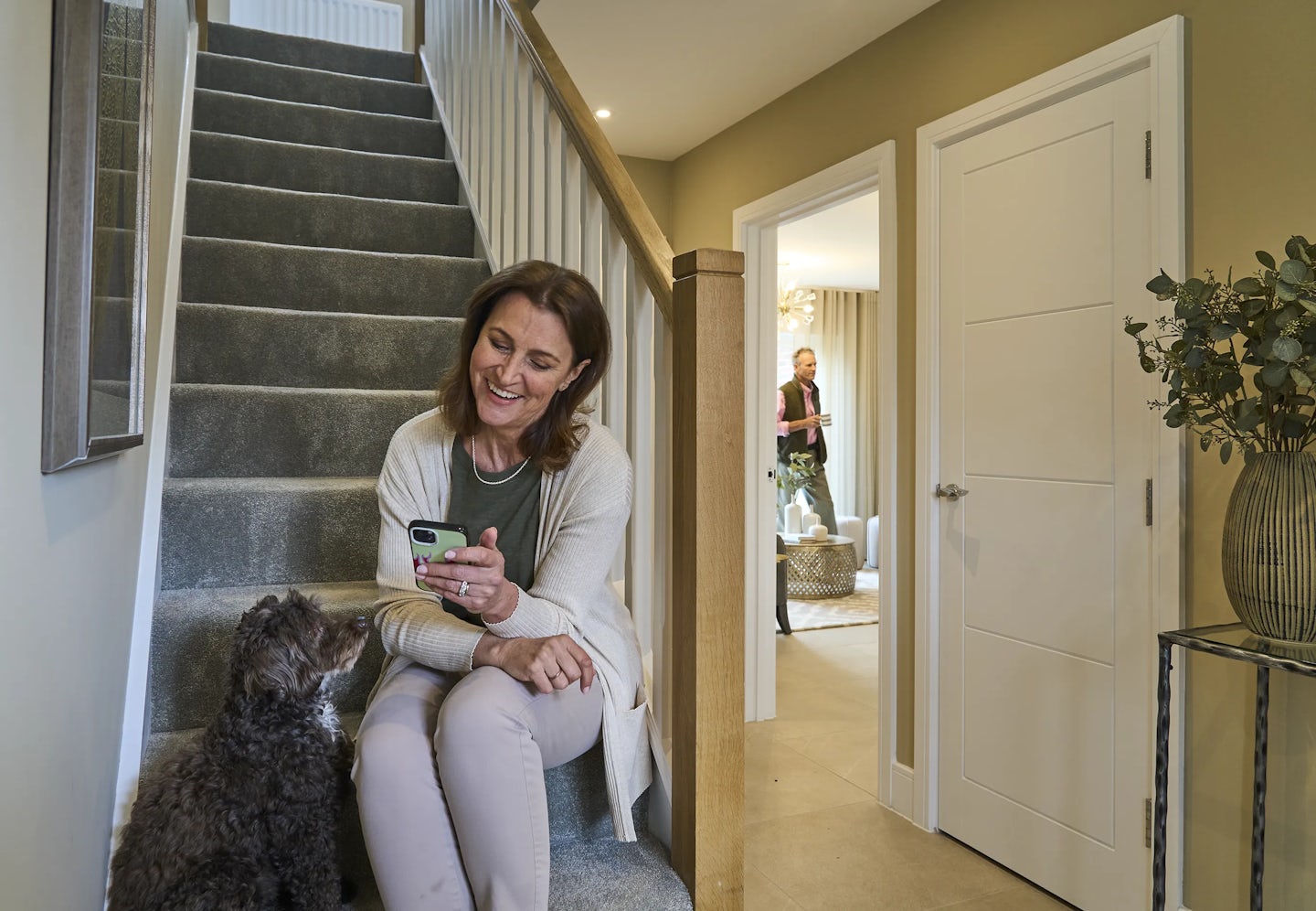 A woman sits on a staircase, holding a smartphone while a dog sits beside her, looking up. The staircase has grey carpet and wooden railings. In the background, a man stands in a room, holding a mug, with a hallway leading to a white door and a small table with a plant.