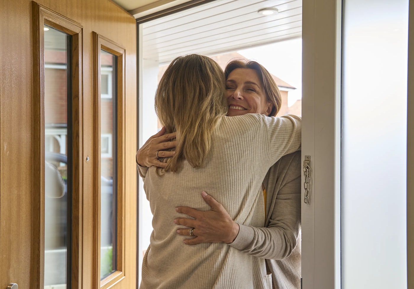 The image shows two people embracing at the entrance of a house. Both individuals are wearing light-coloured sweaters. The scene appears to depict a warm greeting or farewell. The door is open, showing the interior of the house, including a wooden door with glass panels.