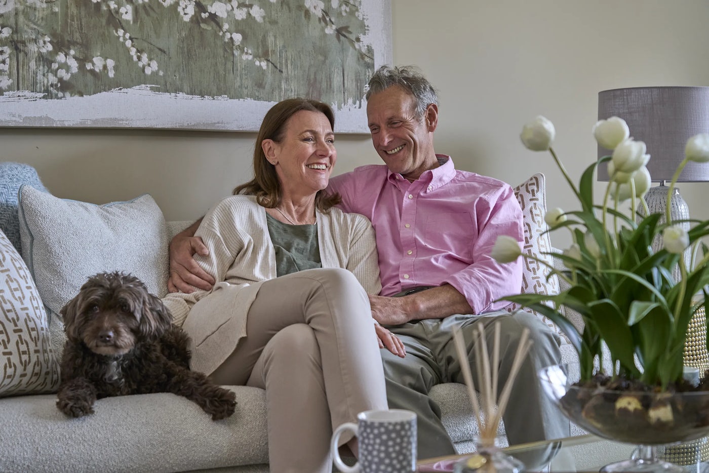 A living room with a downsizer couple sitting on a sofa. The person on the left holds a small dog. The space is decorated in neutral tones, featuring a large window with curtains, a coffee table with books, a plant, and decorative objects.
