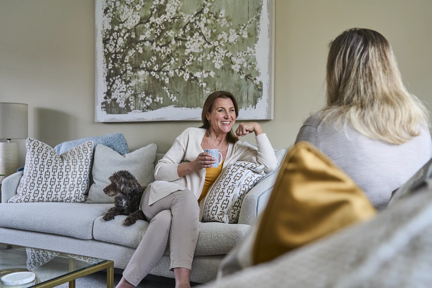 Two women sit on a sofa in a living room, engaged in conversation. One holds a cup while a small dog sits beside them. The room is decorated with patterned pillows and a large piece of floral artwork hanging on the wall behind them.