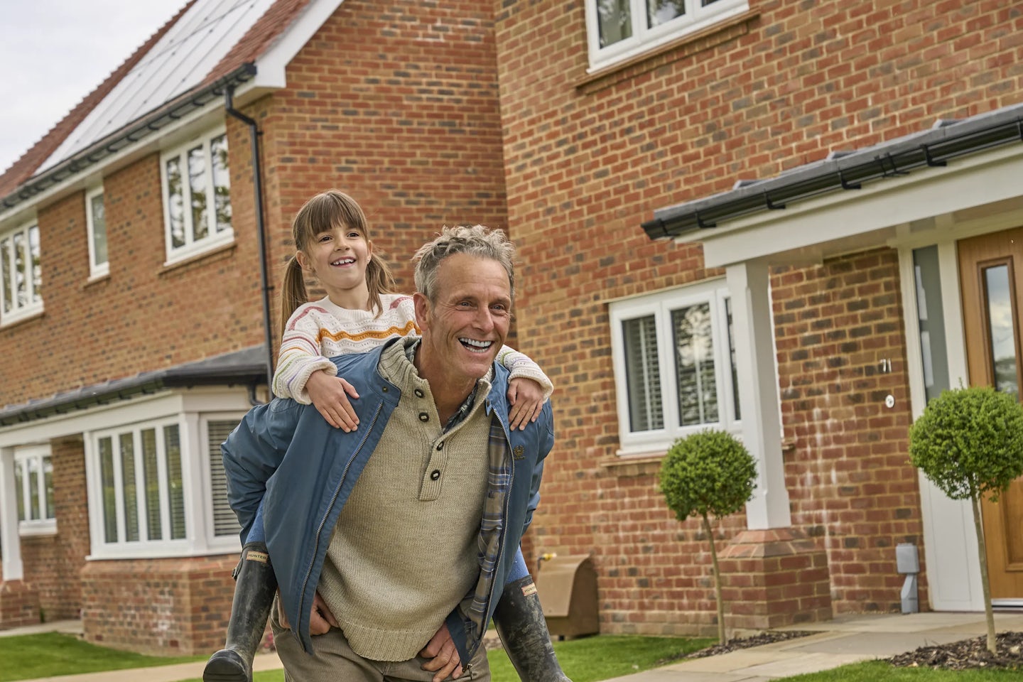 A man carries a young girl on his back outside a brick house. The house features white-framed windows, a wooden front door, and two small trees in front. Solar panels can be seen on the roof of the house next door.