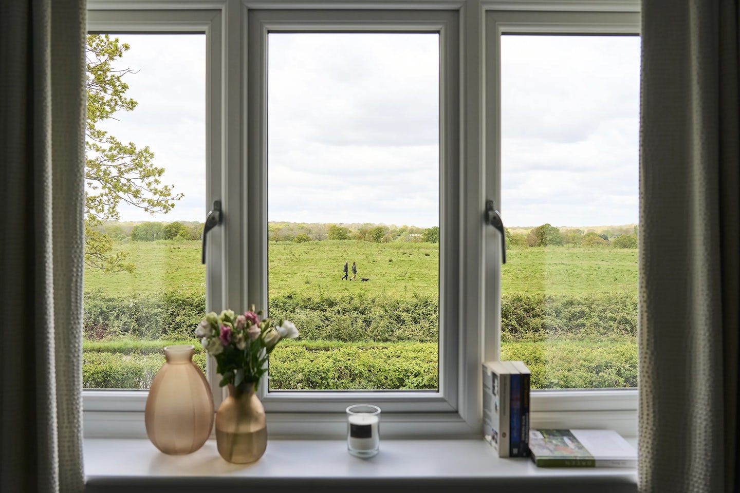 A view through a three-panel window overlooking a lush green field. Two people walk in the distance, adding a quiet human presence to the serene landscape. The window sill is adorned with a vase of flowers, another decorative vase, a candle, and a few books, creating a cosy and thoughtful interior scene.