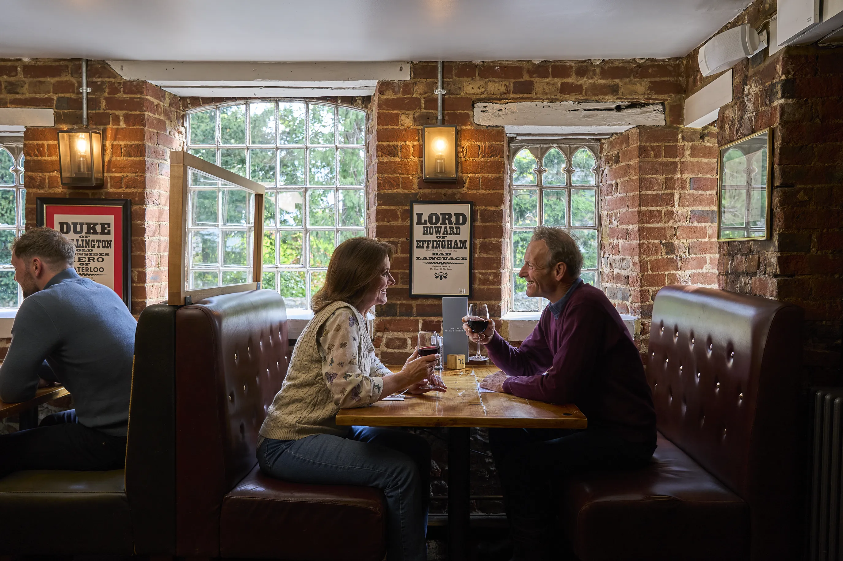Two individuals sit across from each other at a table in a restaurant or pub with exposed brick walls. They are engaged in conversation, holding glasses of red wine. Behind them, framed posters on the wall include text referencing historical figures: "DUKE OF WELLINGTON THE HERO OF WATERLOO" and "LORD HOWARD OF EFFINGHAM." Large windows allow natural light to filter into the space. A man sits in an adjoining booth.