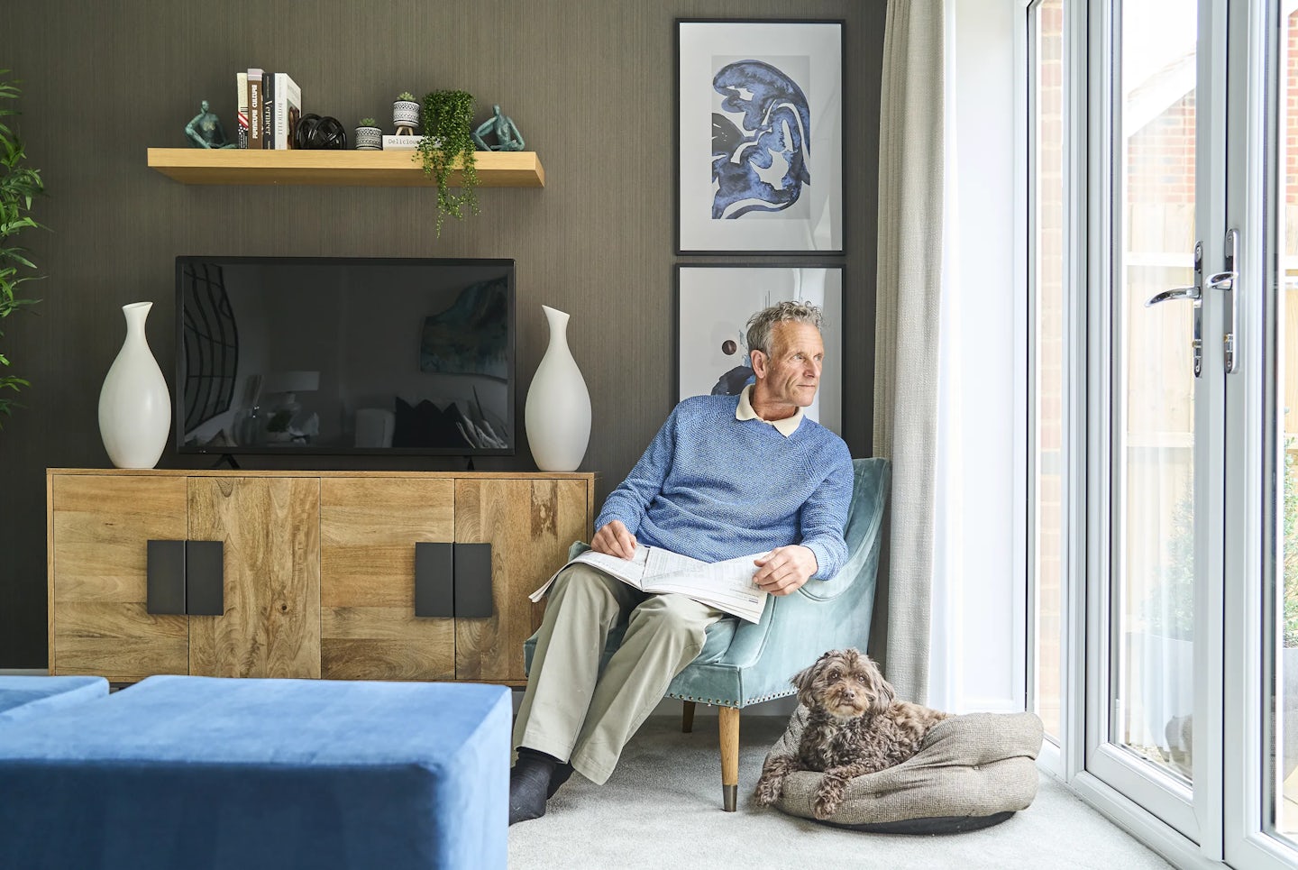 A man wearing a blue sweater and beige pants sits in a chair, reading a newspaper. A small dog lies on a cushion on the floor nearby. The living room features a wooden cabinet with a television on top, flanked by two white vases. Above the television, a wooden shelf holds books, decorative items, and a small plant. Two framed abstract artworks are mounted on the wall behind him. Large glass doors allow natural light into the space.