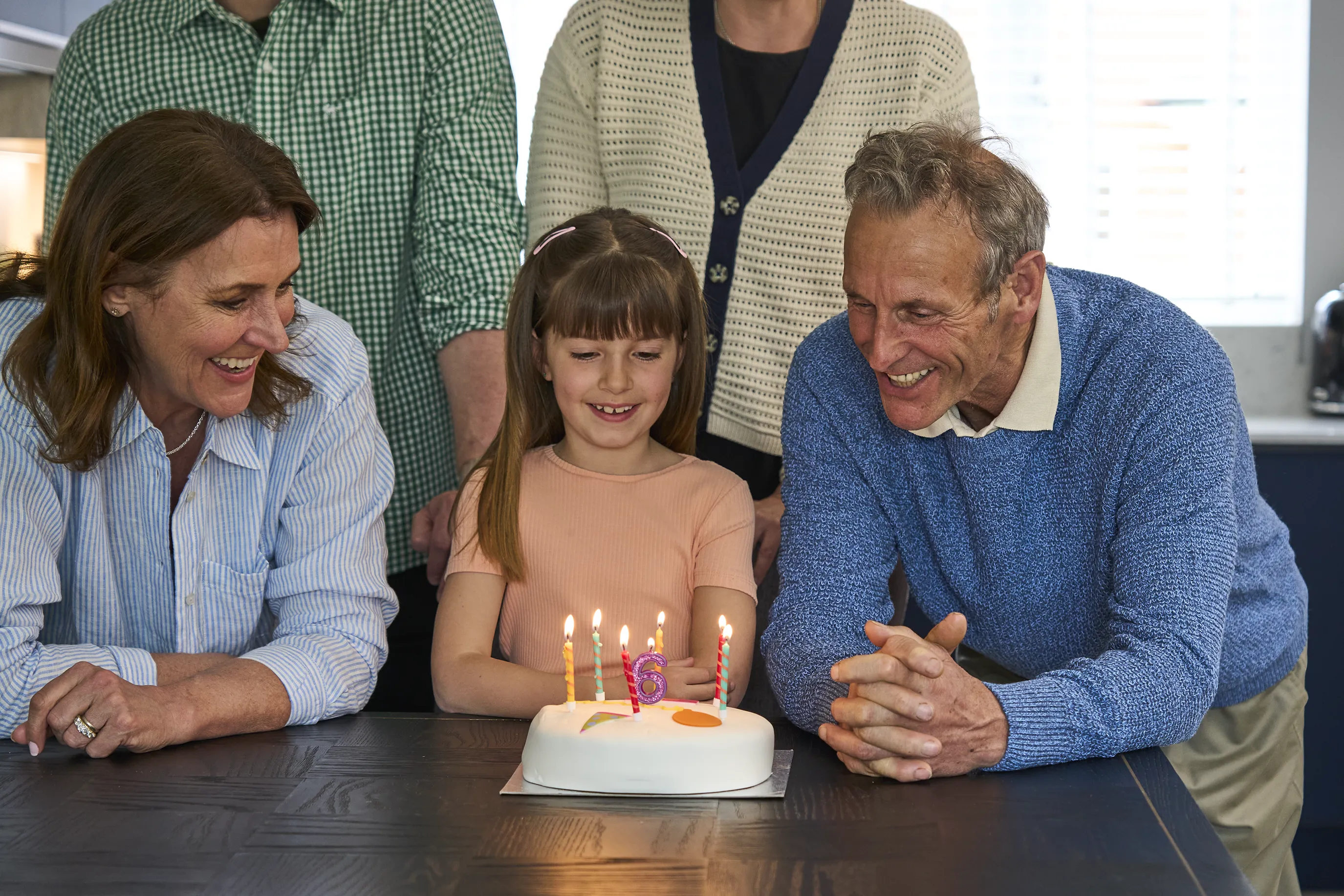 The image depicts a family gathering around a table with a birthday cake that has lit candles. The cake features a number "6" candle, suggesting it's a celebration for a child's sixth birthday.