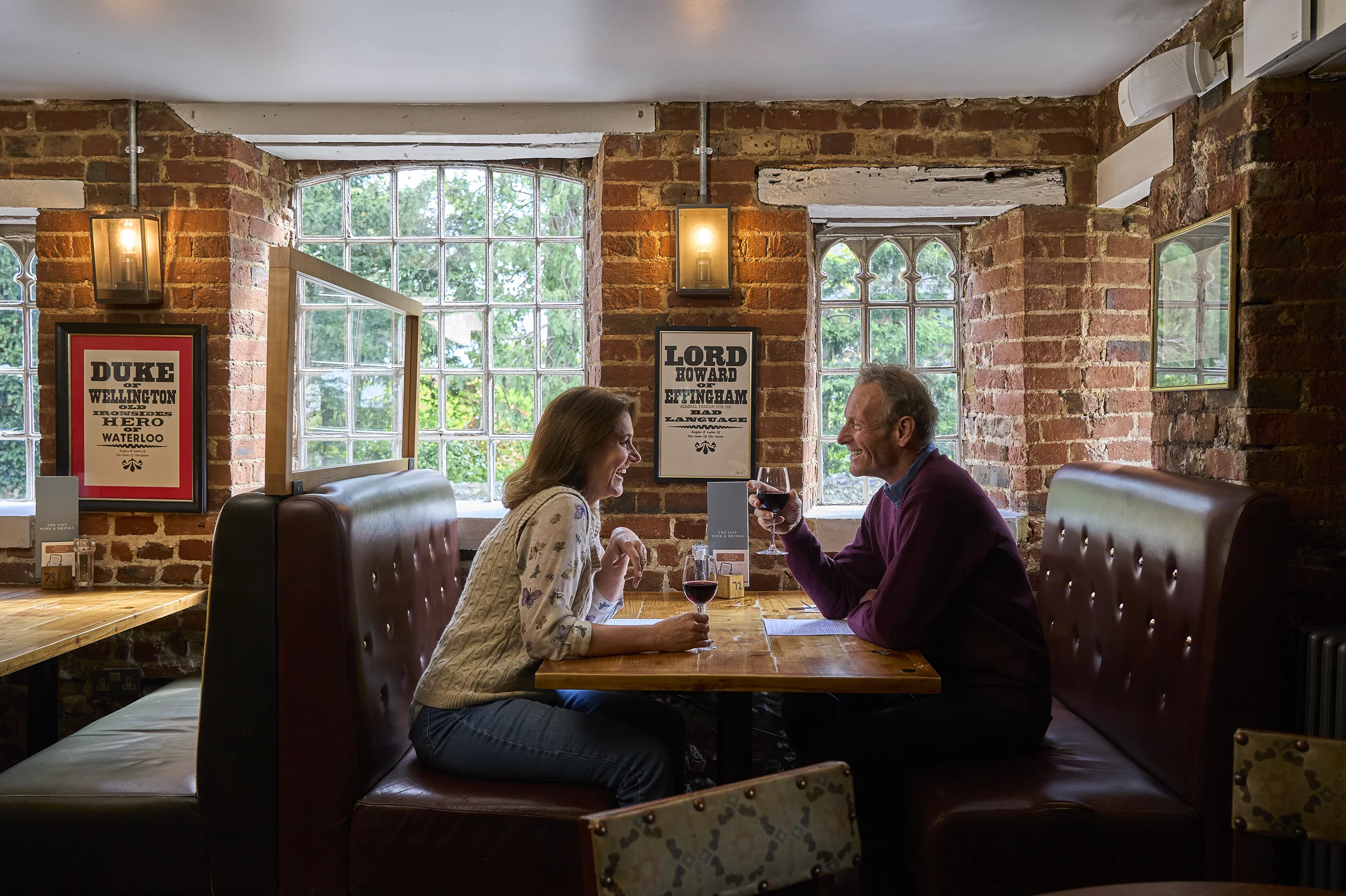 Two individuals sit across from each other at a table in a restaurant or pub with exposed brick walls. They are engaged in conversation, holding glasses of red wine. Behind them, framed posters on the wall include text referencing historical figures: "DUKE OF WELLINGTON THE HERO OF WATERLOO" and "LORD HOWARD OF EFFINGHAM." Large windows allow natural light to filter into the space.