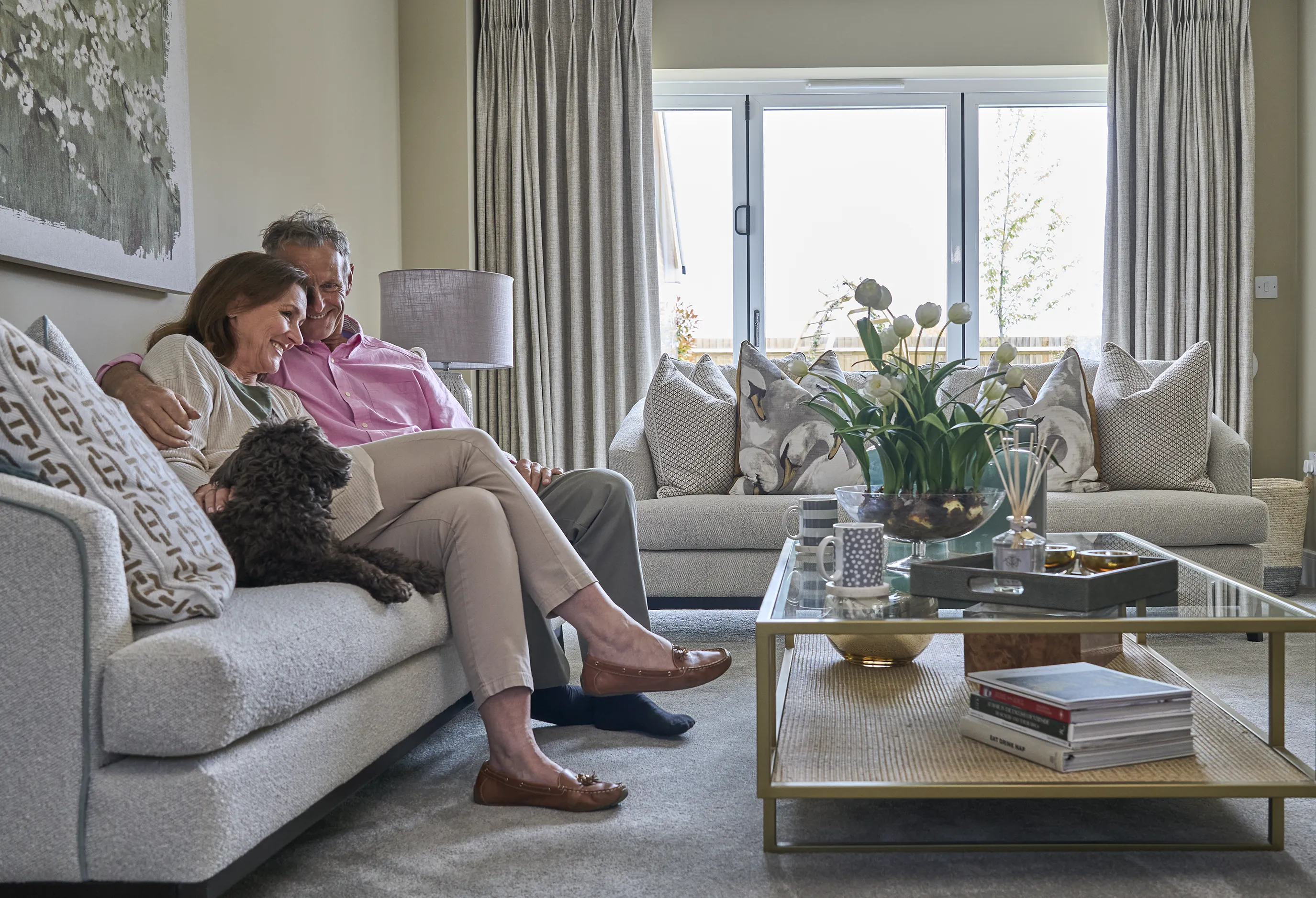 A living room with a downsizer couple sitting on a sofa. The person on the left holds a small dog. The space is decorated in neutral tones, featuring a large window with curtains, a coffee table with books, a plant, and decorative objects. A second sofa in the background is adorned with several pillows.