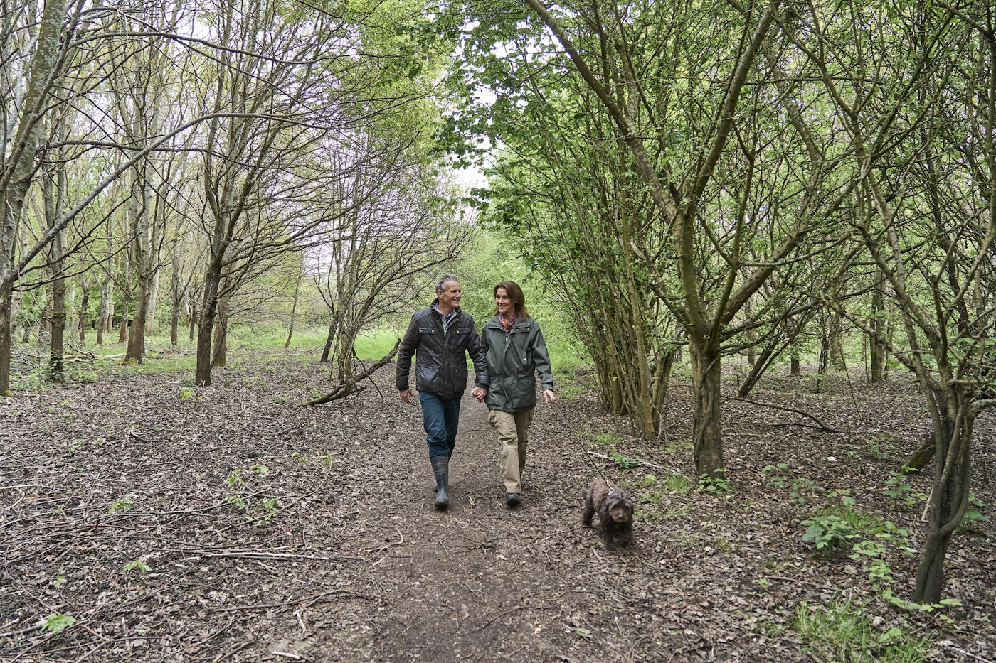 The image shows two people walking along a dirt path in a wooded area. The path is surrounded by trees with green foliage, and the ground is covered with fallen leaves and small plants. The two people are walking side by side, and there is a small dog walking ahead of them.