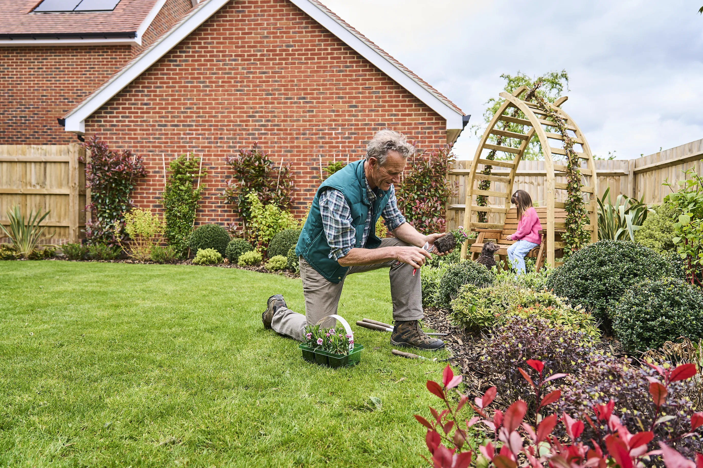 A man kneels on the grass in a garden, planting flowers in a flower bed. He wears a green vest over a plaid shirt and beige pants. A small container of flowers sits beside him. In the background, a young girl sits on a wooden structure, surrounded by plants and bushes. The well-maintained garden features lush greenery, a brick house with a solar panel on the roof, and various plants.