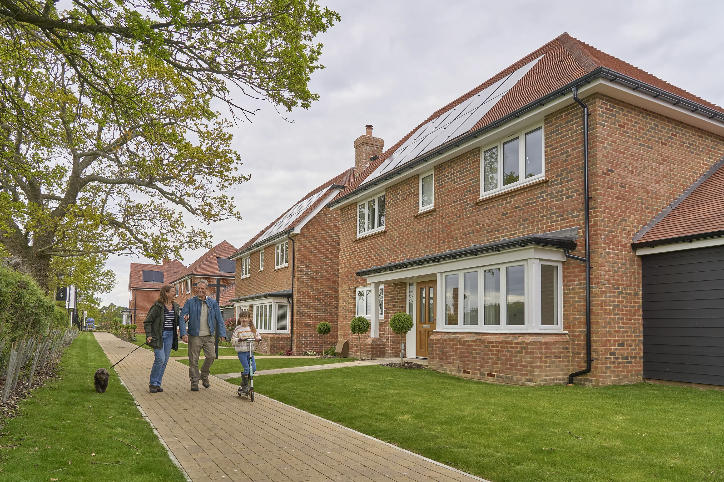 A downsizer couple walks along a paved pathway in a suburban neighborhood, accompanied by a young child and a dog. The row of modern brick houses features well-maintained lawns, and some have solar panels on the roofs. The scene reflects a peaceful residential setting with elements of sustainable living and a family-oriented atmosphere.