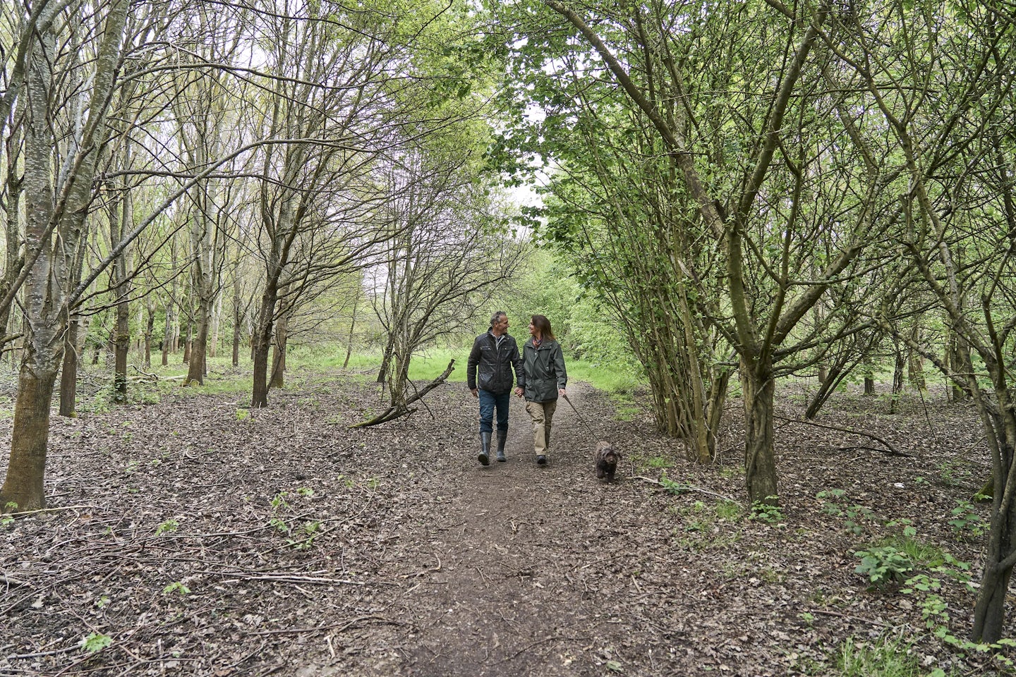 The image shows two people walking along a dirt path in a wooded area. The path is surrounded by trees with green foliage, and the ground is covered with fallen leaves and small plants. The two people are walking side by side, and there is a small dog walking ahead of them.