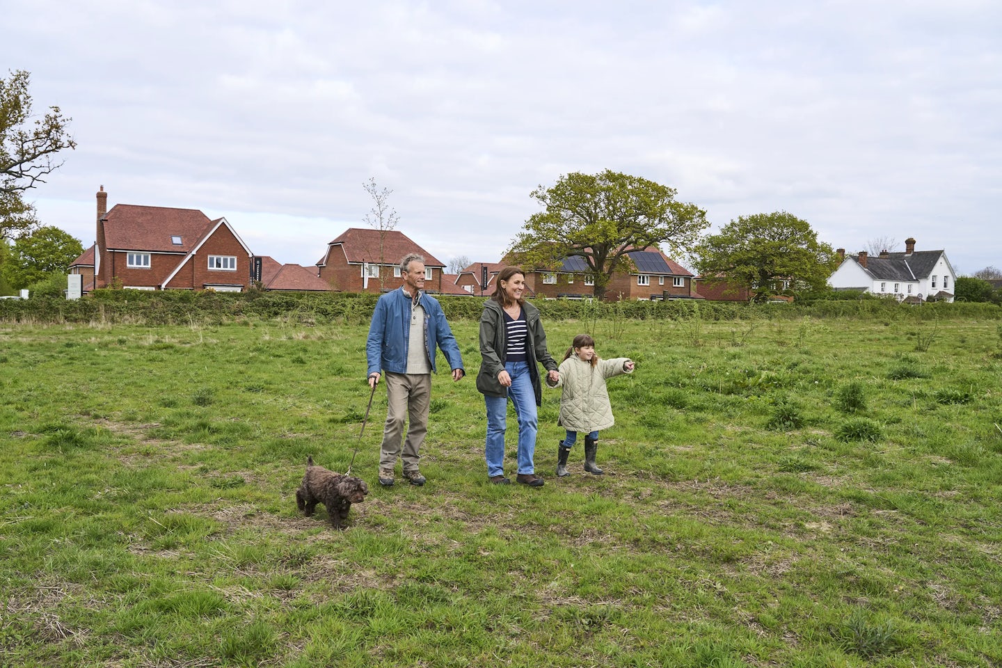 Three people and a dog walk across a grassy field. The group consists of an older man, a middle-aged woman, and a young child. The older man holds the dog's leash. In the background, there are several houses with red roofs and scattered trees. The setting appears to be suburban or rural, with a relaxed and peaceful atmosphere.