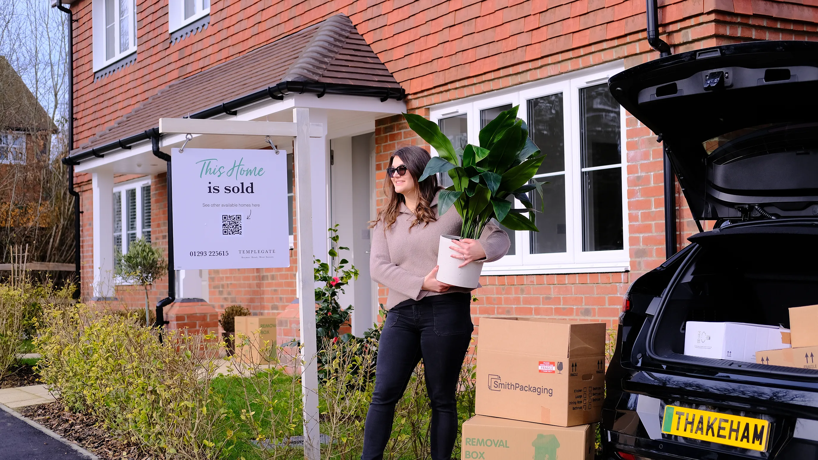 Woman carrying a plant from the boot of a black car in front of a new home. The ground floor is brick and the first floor is clad in shingle tile. In front of the house is a 'This home is sold' sign.
