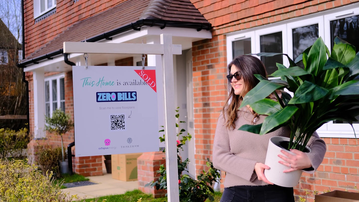 A woman holds a plan next to a sign that says 'This home is sold', in front of a new build brick home. The sign also indicates the home is Zero Bills by Octopus Energy.