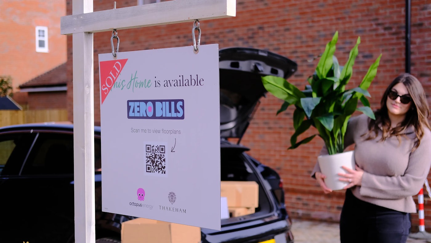 A woman carries a plant from the open boot of  black car. In front in focus is a sign that says 'This home is sold', the sign also indicates the home is Zero Bills by Octopus Energy.