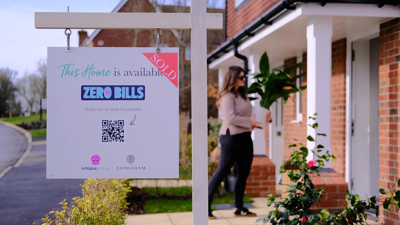 A woman carries a plant into a new build brick home. In focus is a sign that reads 'This home is sold'. The sign also indicates the home is Zero Bills by Octopus Energy.