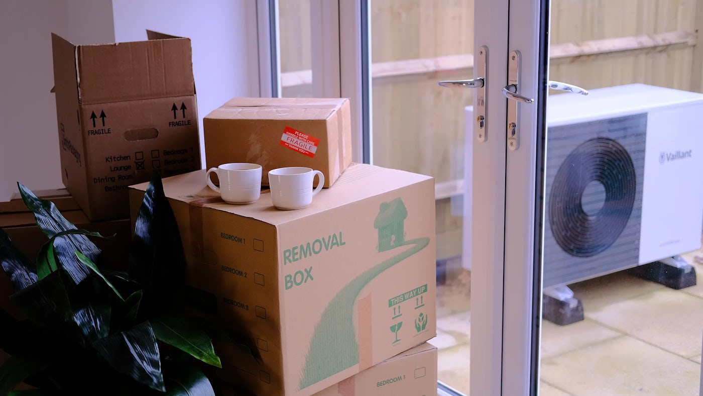 Removal boxes with fragile stickers are stacked in front of patio doors. Two cups of tea are resting on a box. Outside a heat pump sits against a fence.