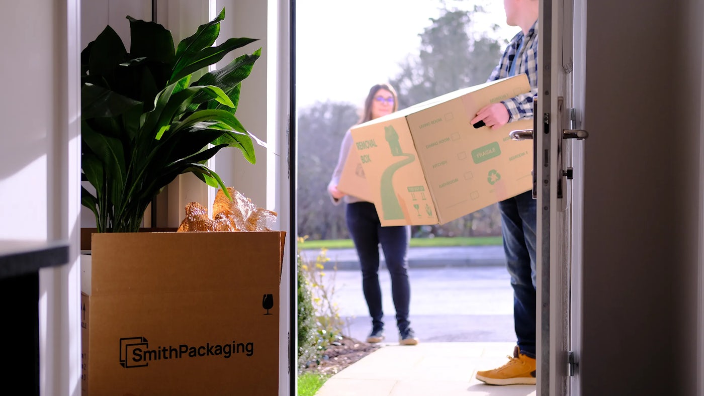 A couple carry boxes in the front door of their new home. Boxes and plants are on the floor inside the door.