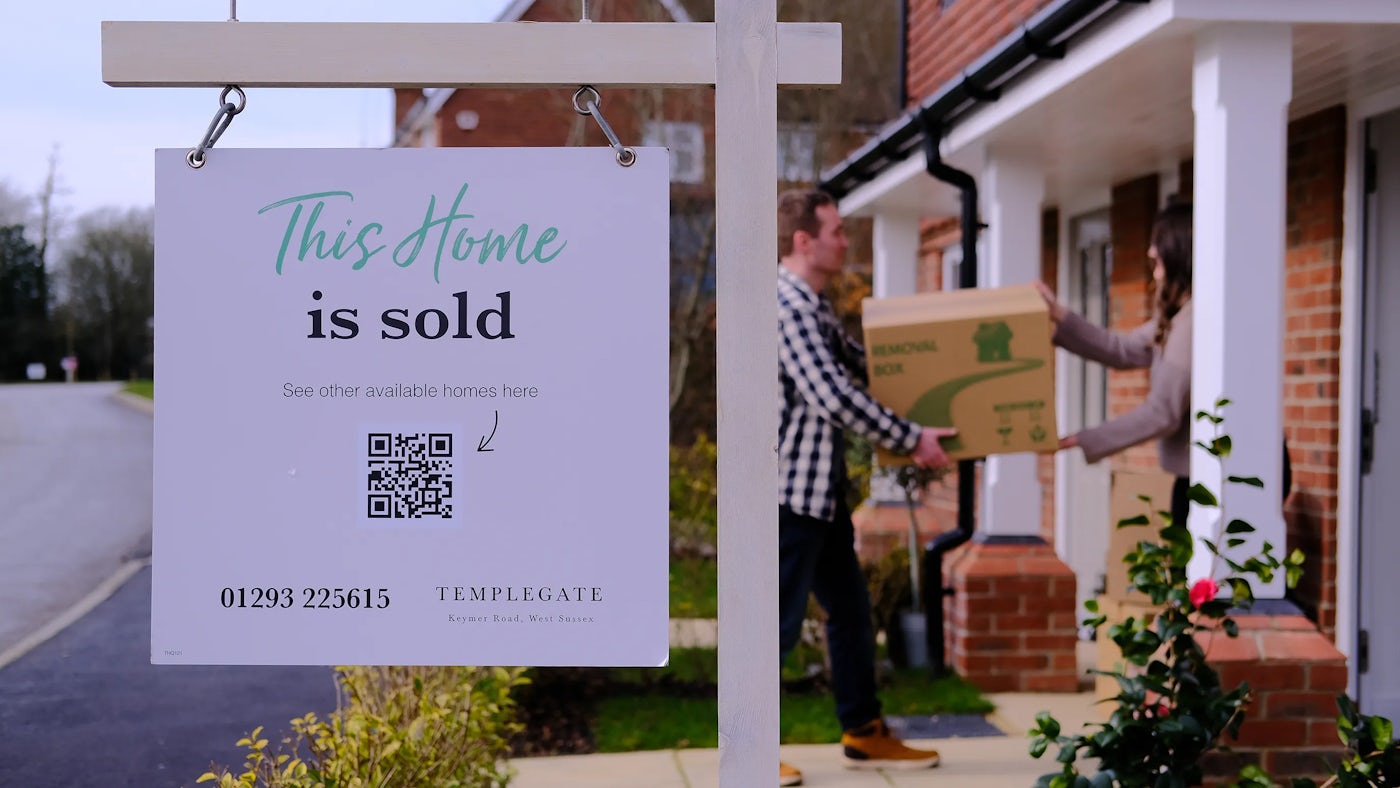 A man passes a box to woman in the doorway of a new build home. In front in focus is a sign that reads 'This home is sold'.