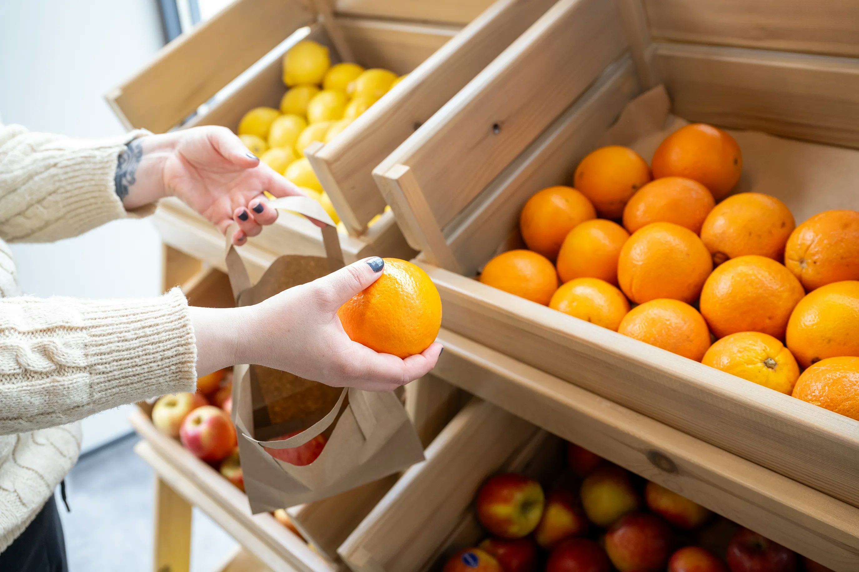 A person is holding a ripe orange in one hand while reaching for a paper bag with the other hand. Behind them, wooden crates filled with oranges are visible, showcasing a vibrant array of fresh fruit, while a lower crate contains red apples. The setting is a community owned shop.