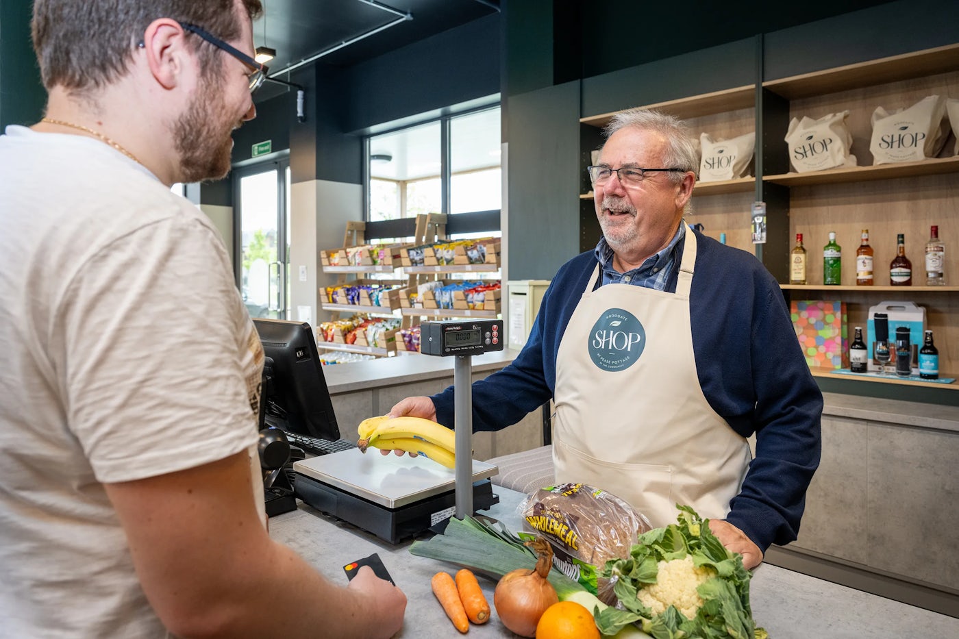 A man standing in front of a counter of a community owned shop with a selection of vegetables and fruit.