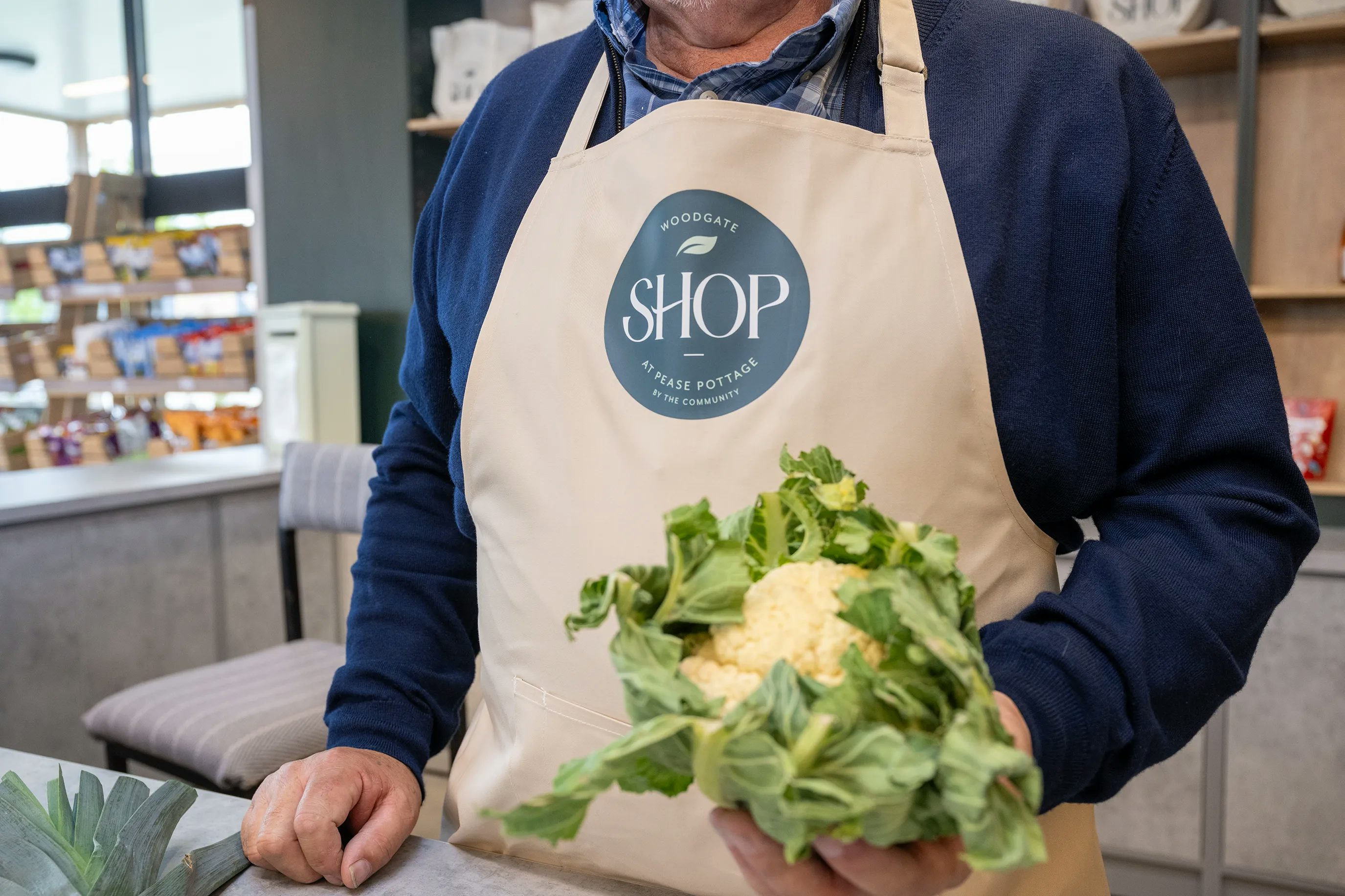 A person wearing a beige apron with the logo "Woodgate Shop" stands in a store holding a fresh cauliflower surrounded by leafy greens. The background features neatly arranged shelves with various grocery items, emphasizing a community-focused shopping experience.
