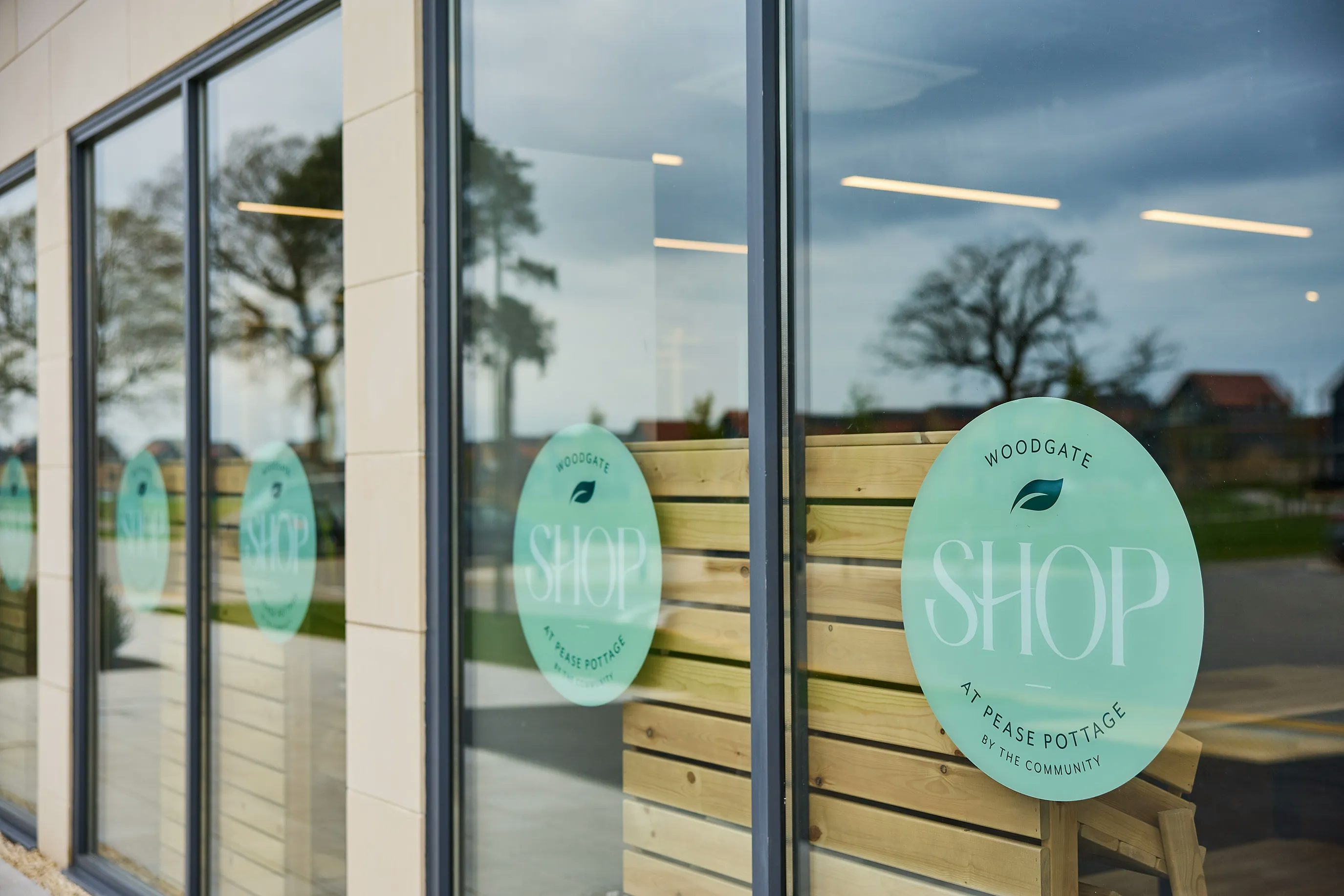 A shop exterior with large glass windows. The windows display circular light green stickers with white text reading "WOODGATE SHOP AT PEASE POTTAGE BY THE COMMUNITY." Reflections in the glass reveal trees and buildings in the background.