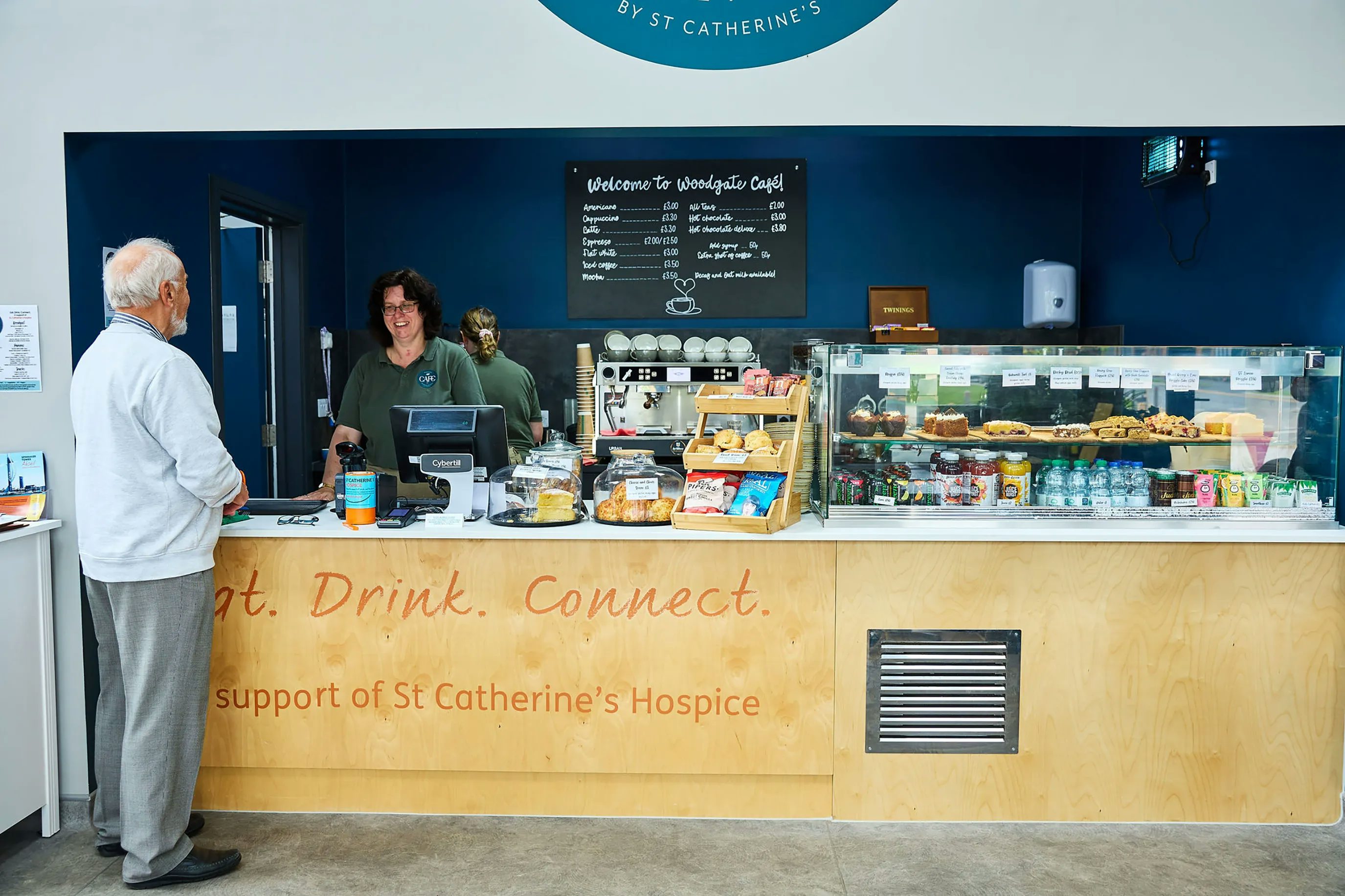 A man stands at the counter of the Woodgate cafe, being served by a lady behind the counter. A range of pastries, cakes and snacks are on display.