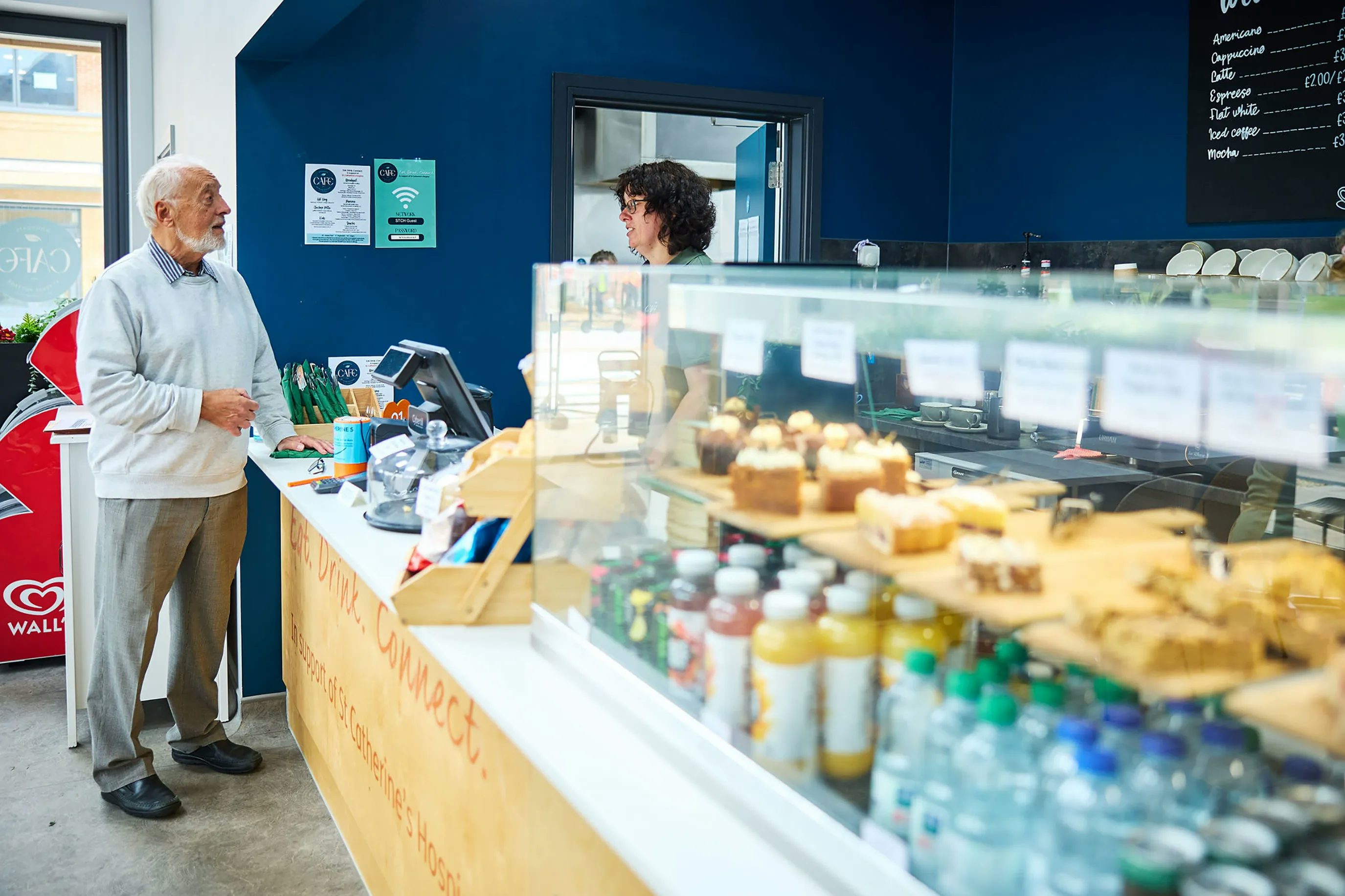 A modern café at Woodgate with a clean and well-organized design. A customer stands at the counter while a staff member is behind it. The counter displays bottled drinks, water bottles, and an assortment of pastries and cakes. A menu board on the wall lists various items with prices.