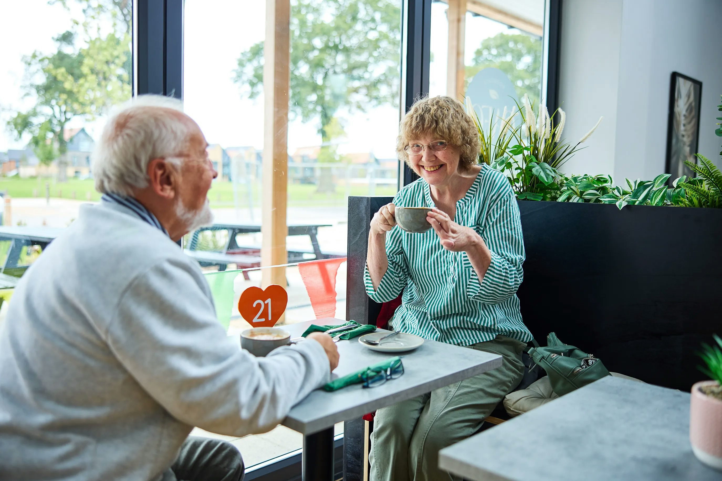 Two people sitting at a table in the Woodgate café. The table has a heart-shaped sign with the number 21, green napkins, cutlery, and two cups—one being held by a woman. Large windows behind them reveal trees and a village green, with plants on a ledge near the seating area.