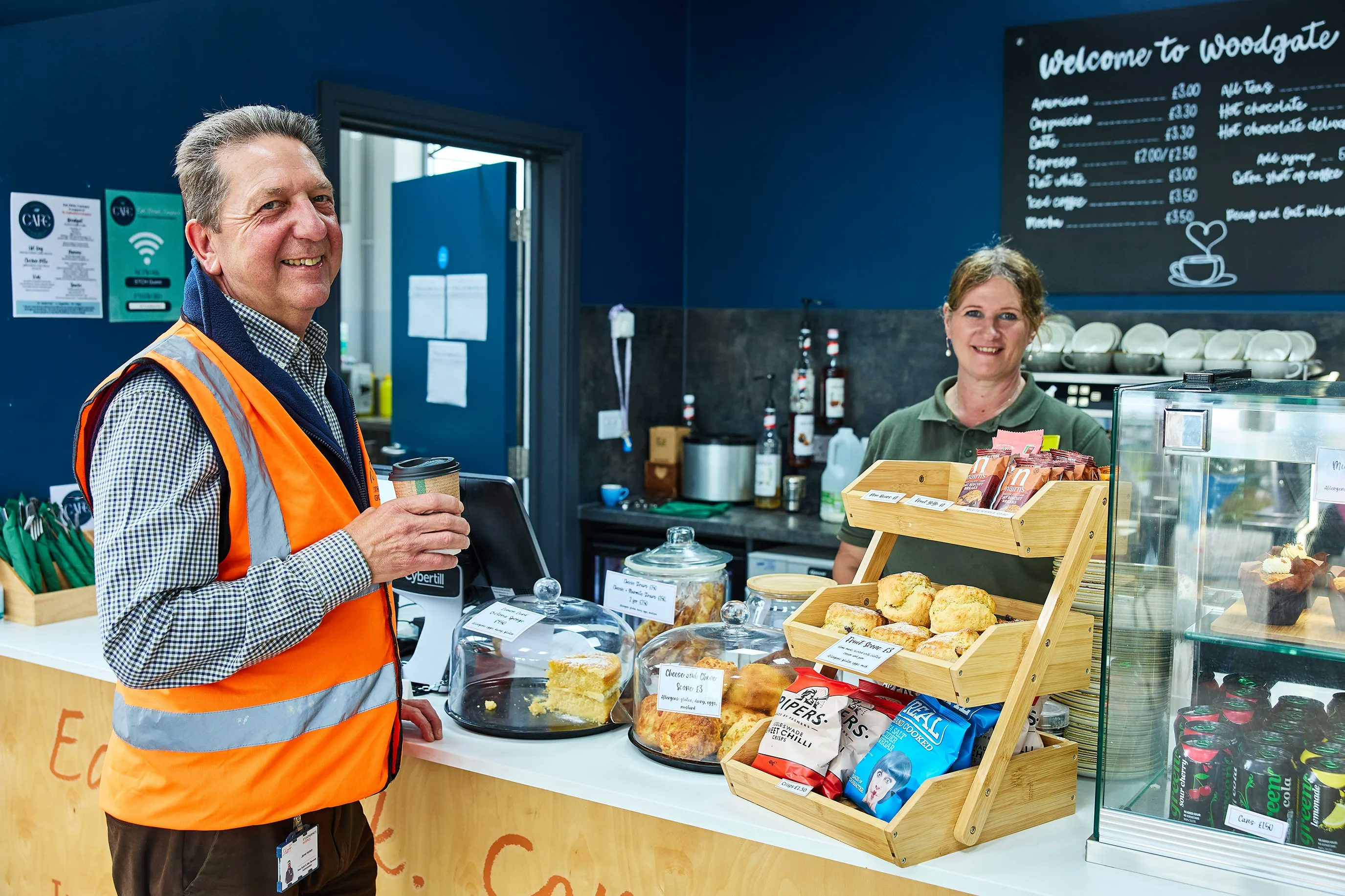 A man in an orange high visibility vest stands with a takeaway coffee cup in front of the counter of the Woodgate Cafe. A lady behind the camera smiles. Pastries, crisps and drinks are on display.