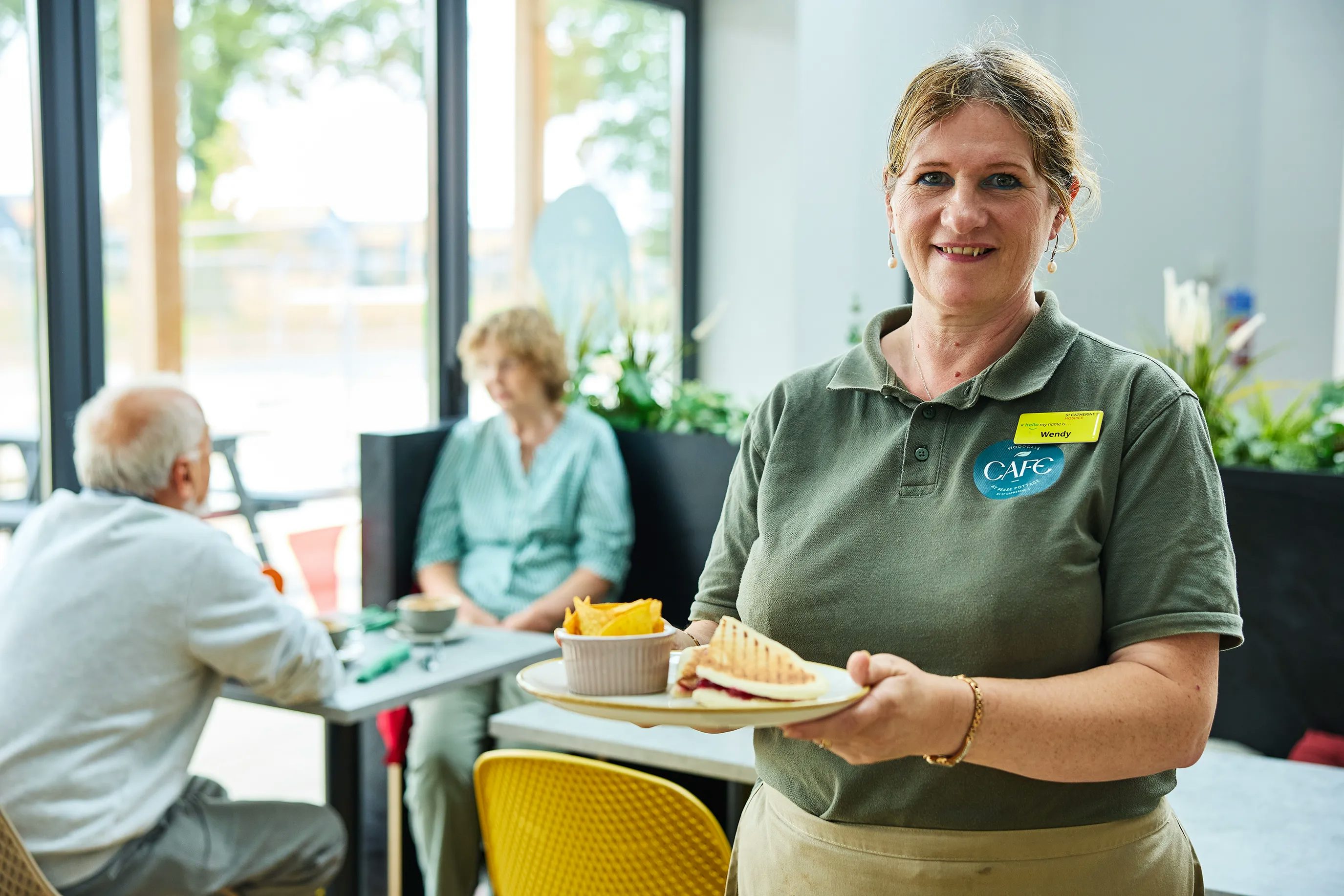 A café employee wearing a green polo shirt with a name tag and a "CAFÉ" logo holds a plate with a sandwich and fries. In the background, two people sit at a table, engaged in conversation, with large windows and indoor plants visible. The setting suggests a casual dining environment.