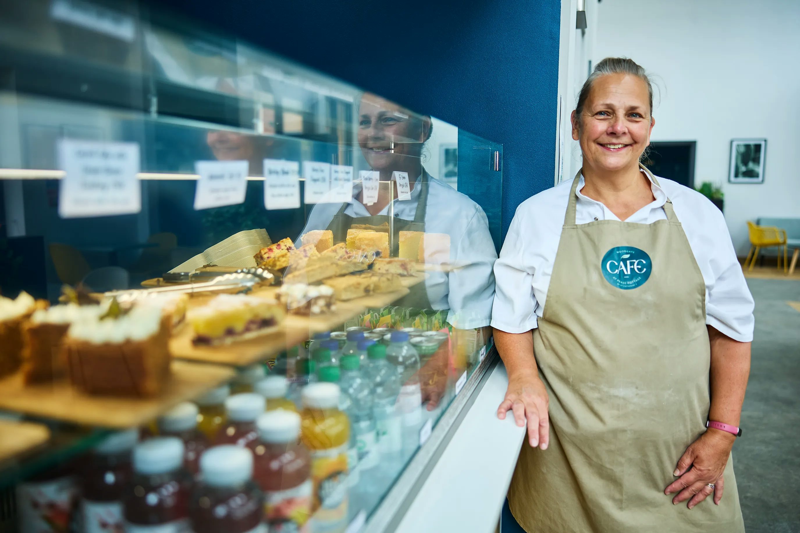 A smiling woman in a white polo shirt and beige apron stands next to a display case filled with various cakes and pastries, in a bright cafe environment.