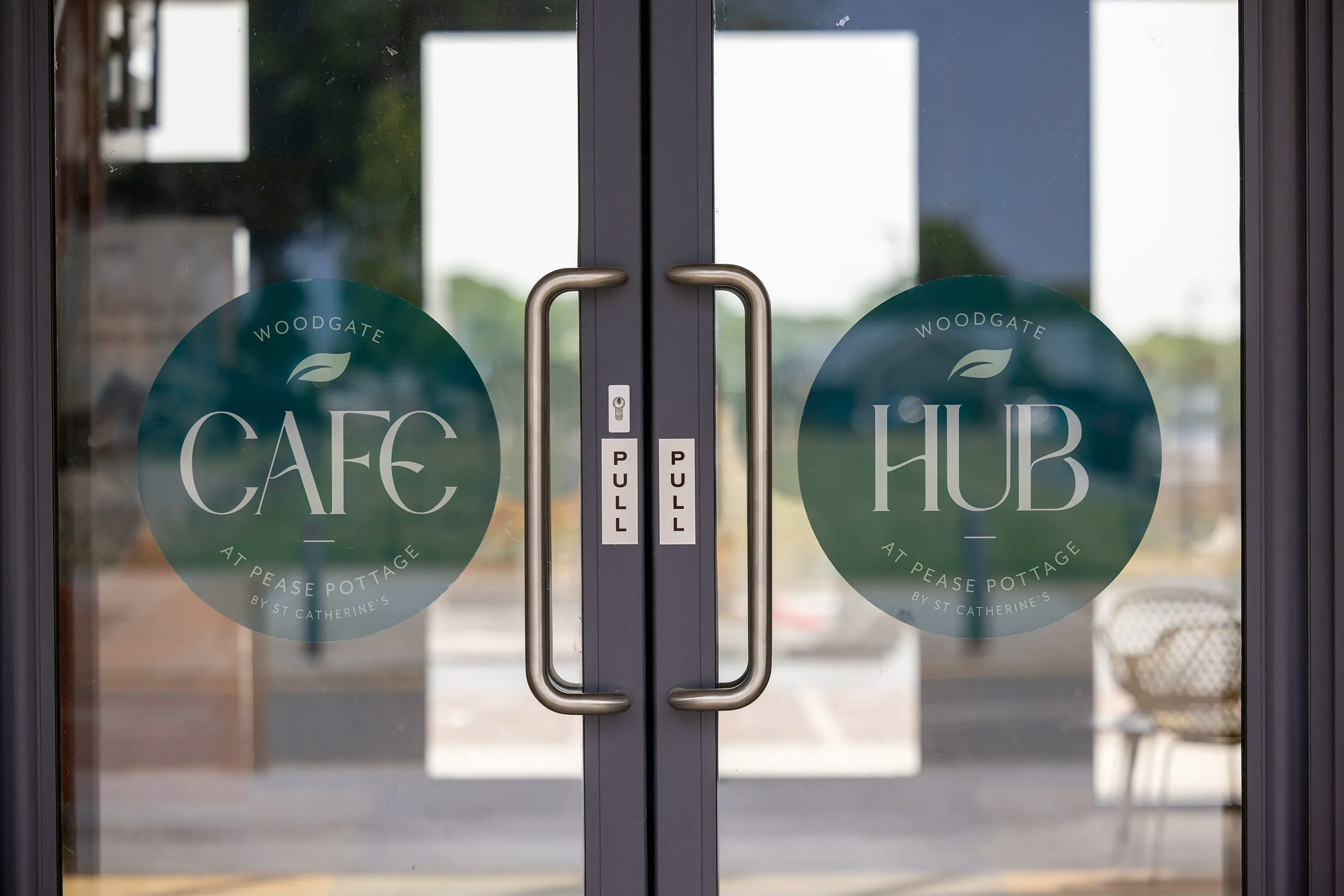 Double glass doors featuring circular signage for "CAFE" and "HUB" at Woodgate, Pease Pottage, with pull handles and the word "PULL" displayed on both sides.