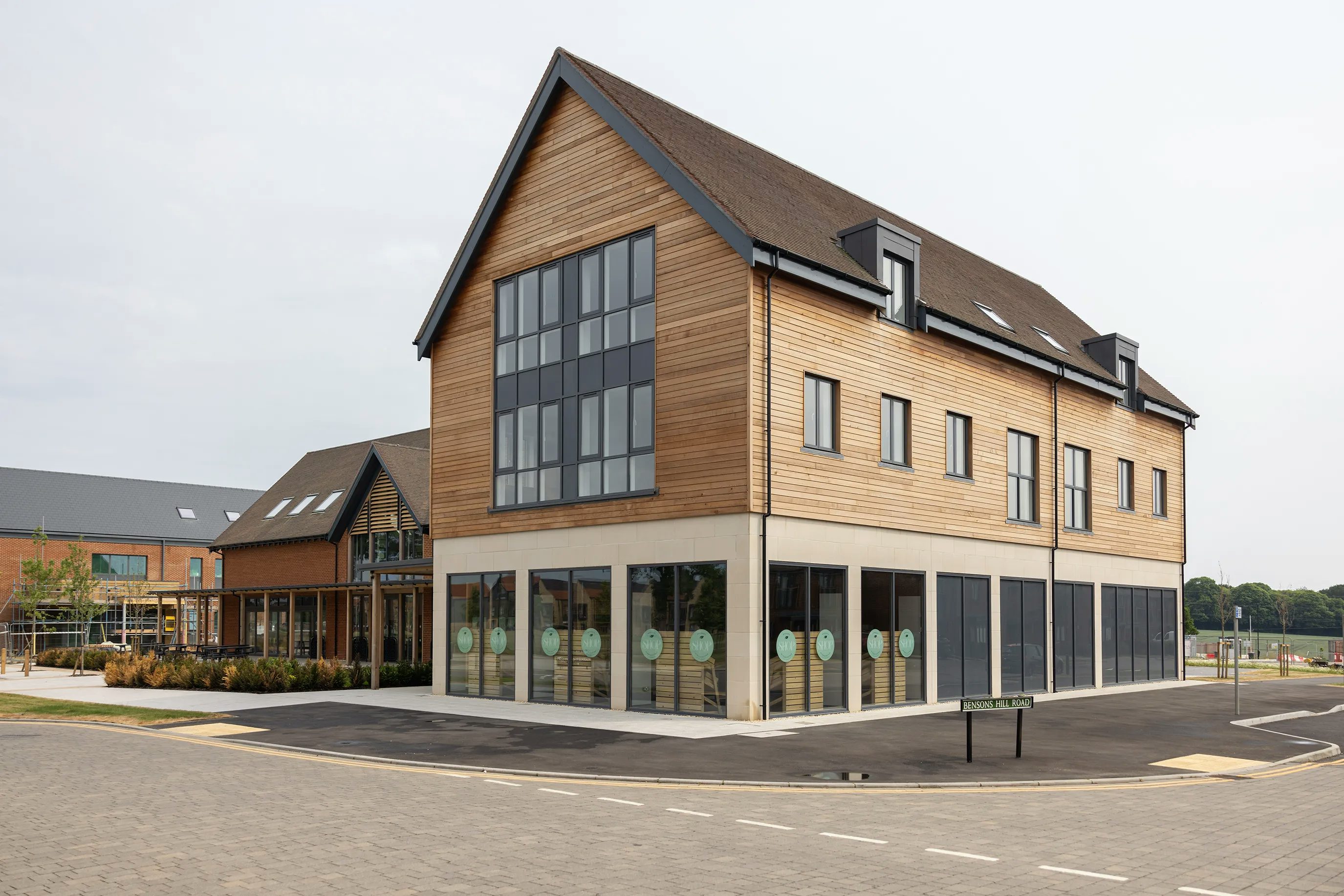 A modern commercial building hold the Woodgate Hub, the Woodgate café and the Woodgate Shop. It has a wooden façade and large glass windows, featuring a pitched roof. The building is located on a paved corner with a sign reading "Foxglove Place," surrounded by similar structures and greenery.