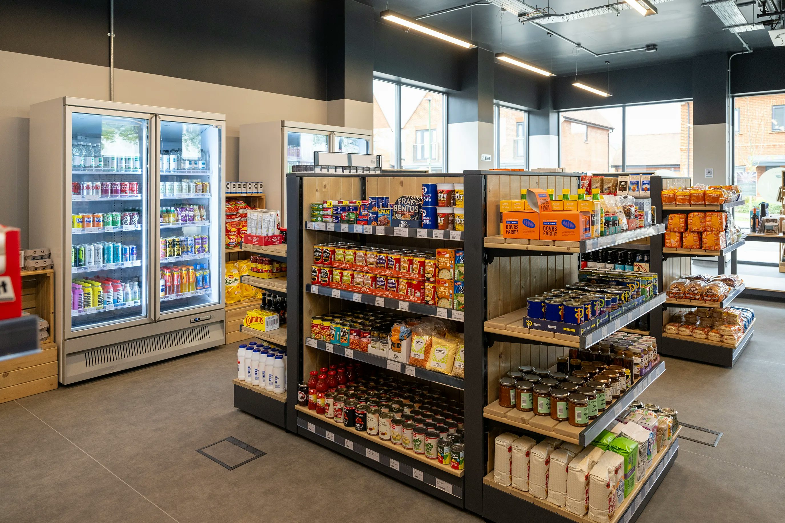 A community owned shop interior with shelves stocked with snacks, canned goods, and beverages. Refrigerated sections on the left display drinks, while the center and right shelves hold various grocery items. Large windows allow natural light to illuminate the organized, clean space