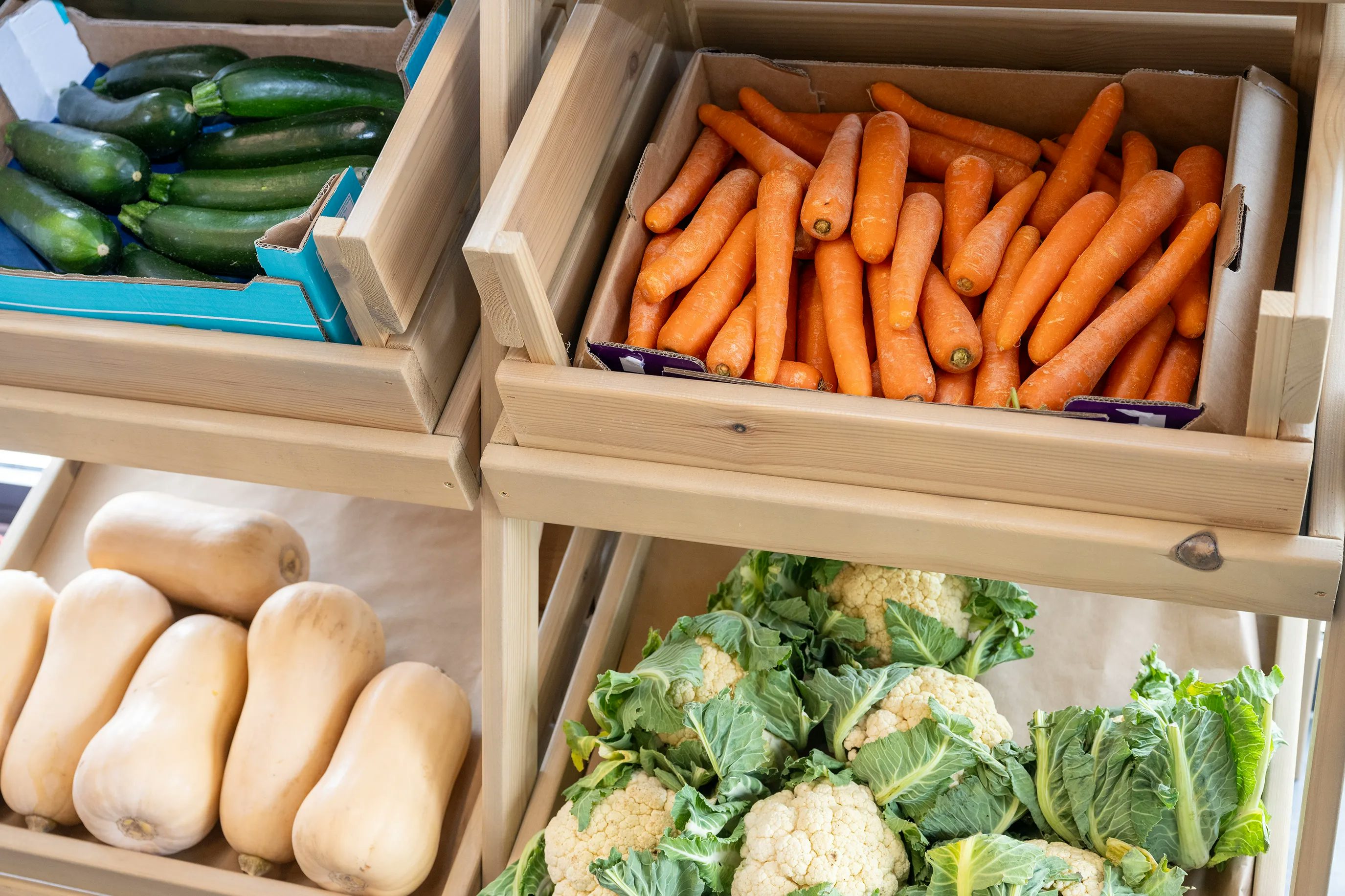 A display of fresh vegetables in a market or grocery store, featuring zucchinis in a blue box on the top left, carrots in a wooden crate on the top right, and butternut squashes and cauliflowers on the lower shelf. The produce is neatly arranged, highlighting its freshness and variety.