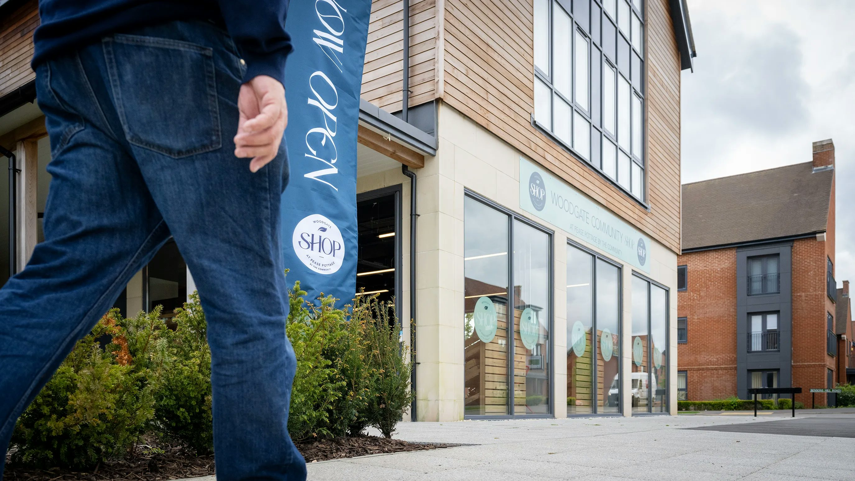 A person walks past a modern building with large glass windows. A blue banner on the exterior reads "NOW OPEN" and "SHOP," indicating a retail space. Above the entrance, a sign reads "WOODCOTE COMMUNITY HUB."