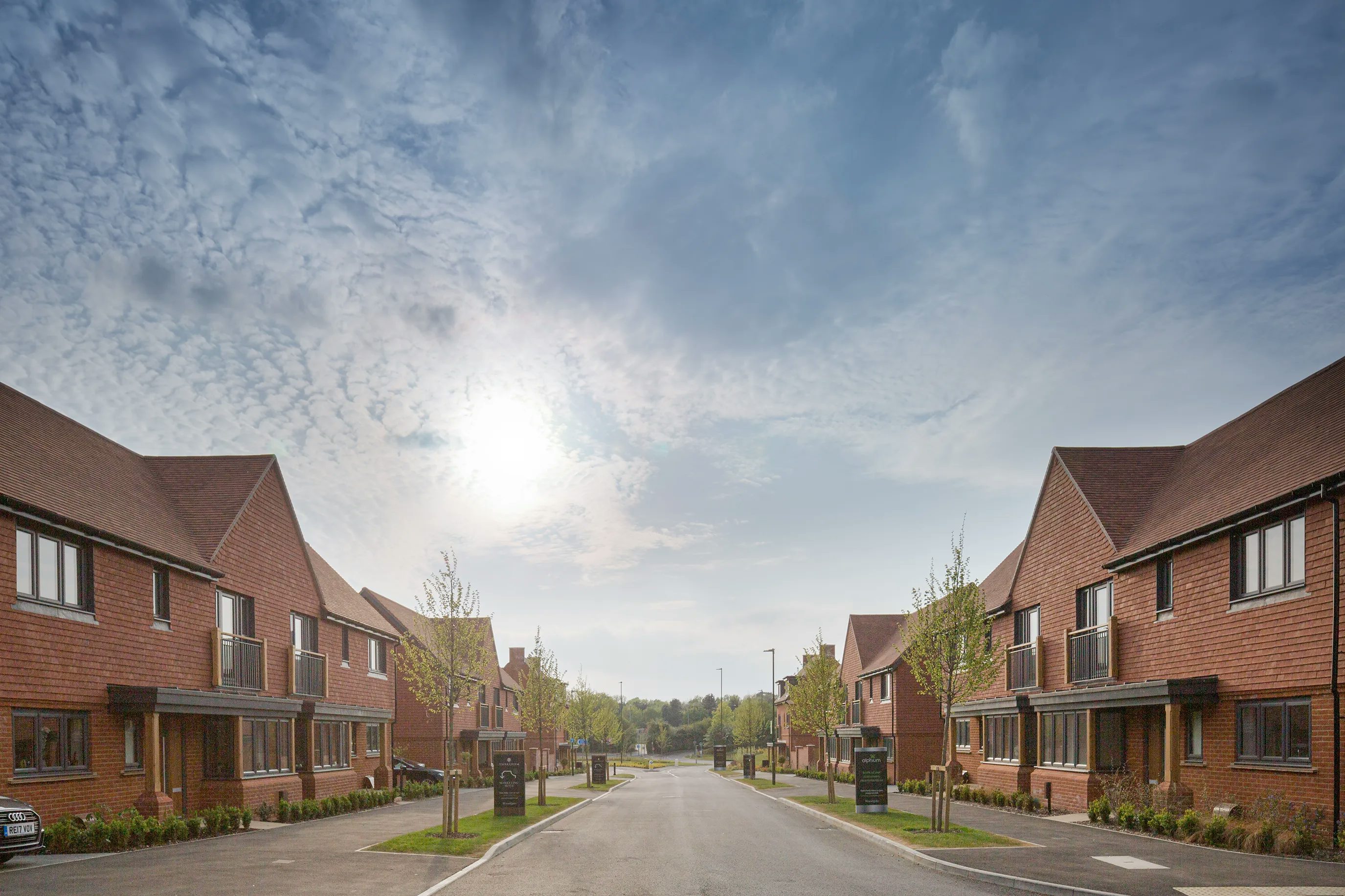 View down the main street into Woodgate at Pease Pottage towards the entrance roundabout. An afternoon sun sits behind scattered clouds.