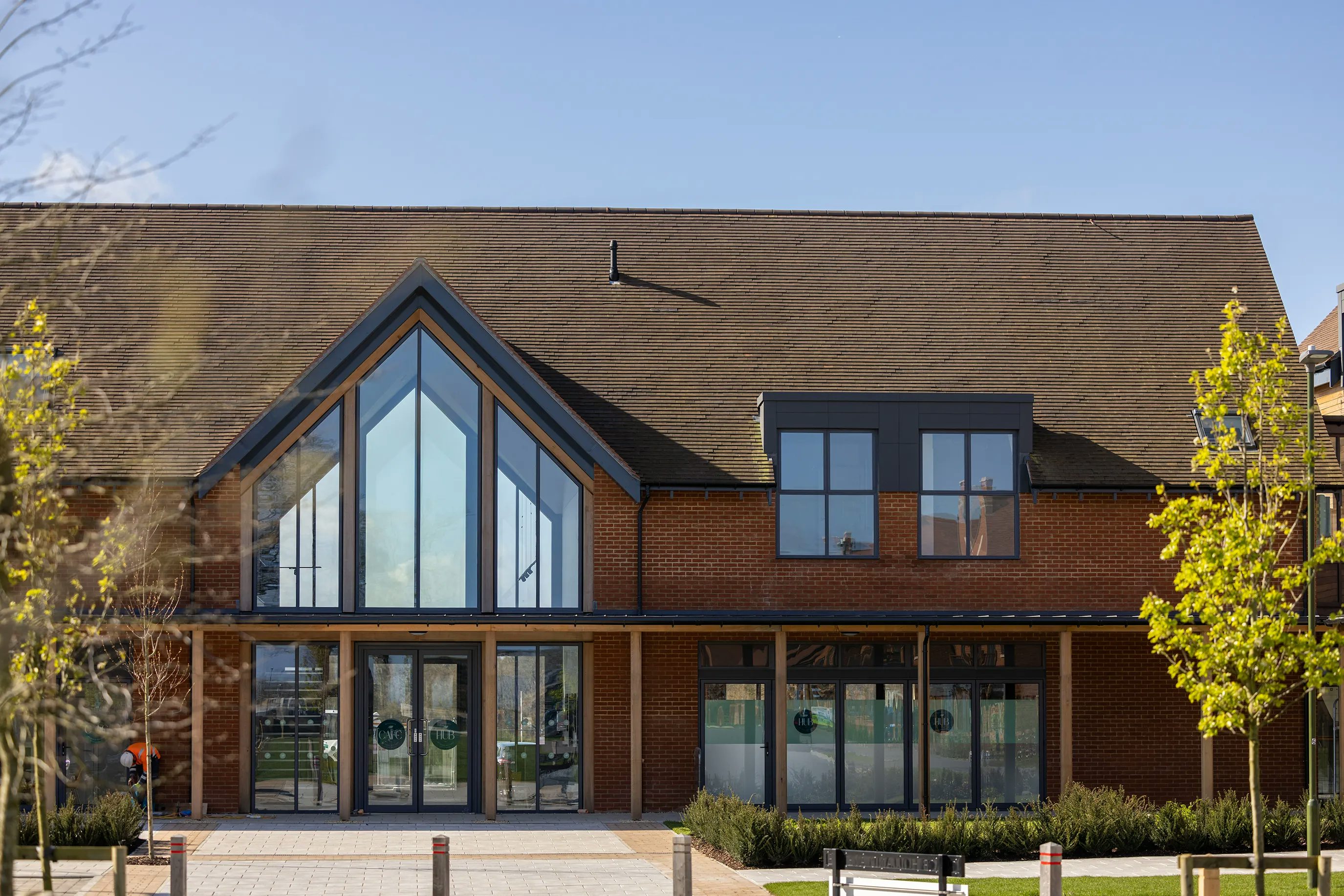 A modern, symmetrical brick building with a triangular roof and expansive glass frontage, featuring two sets of double glass doors marked with circular logos. The building is set within a neatly paved area bordered by bollards, landscaped greenery, and young trees, all under a clear blue sky suggesting a sunny, welcoming atmosphere.