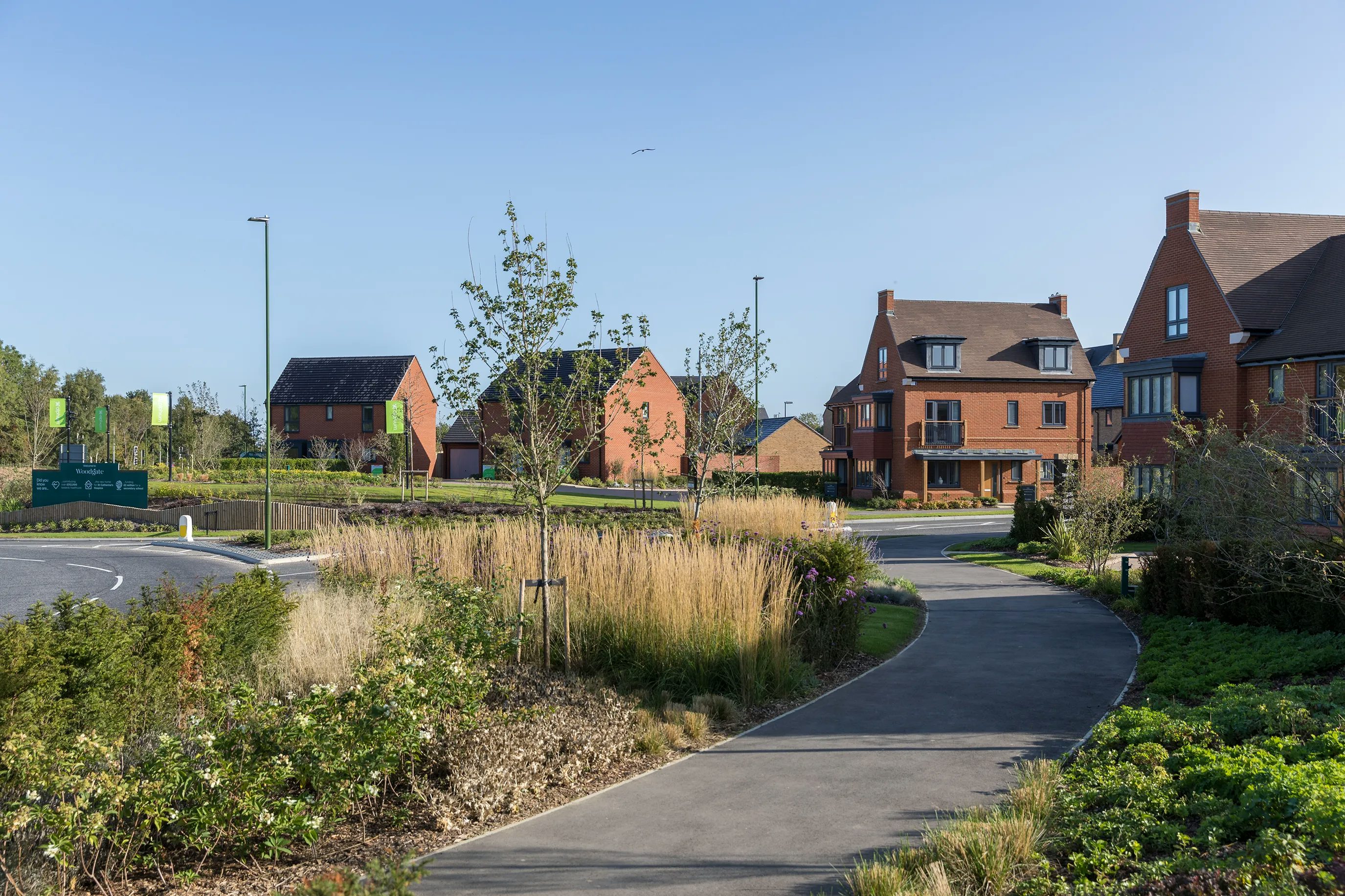 Side view of the main entrance to Woodgate at Pease Pottage. Brick houses sit beside a roundabout. A path to the right hand side is lined by bushes and planting.