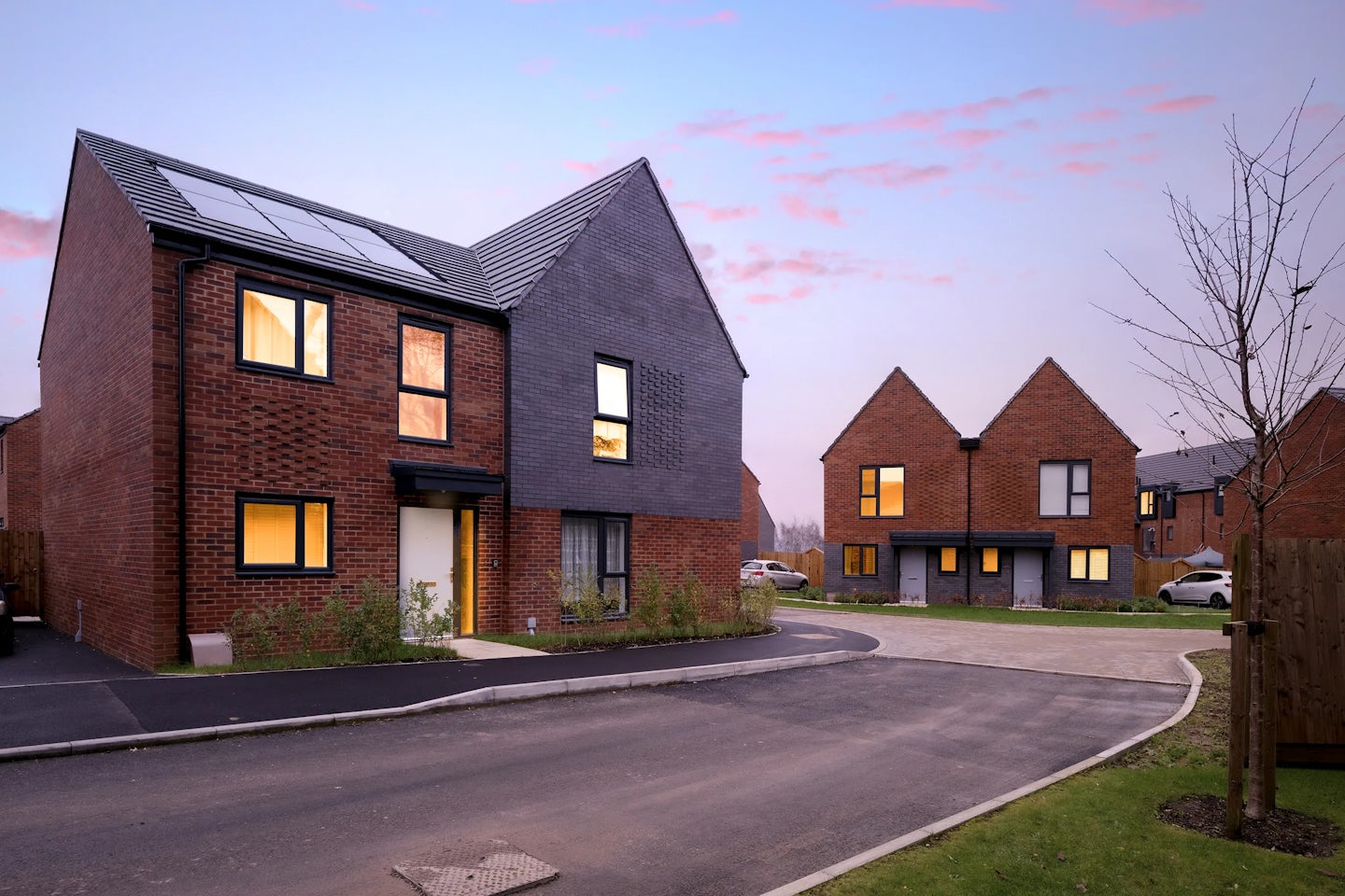 A street of starter homes for first time buyers, in light and dark brick. It's dusk with a purple sky and a few pink clouds. Lights are on in the windows.